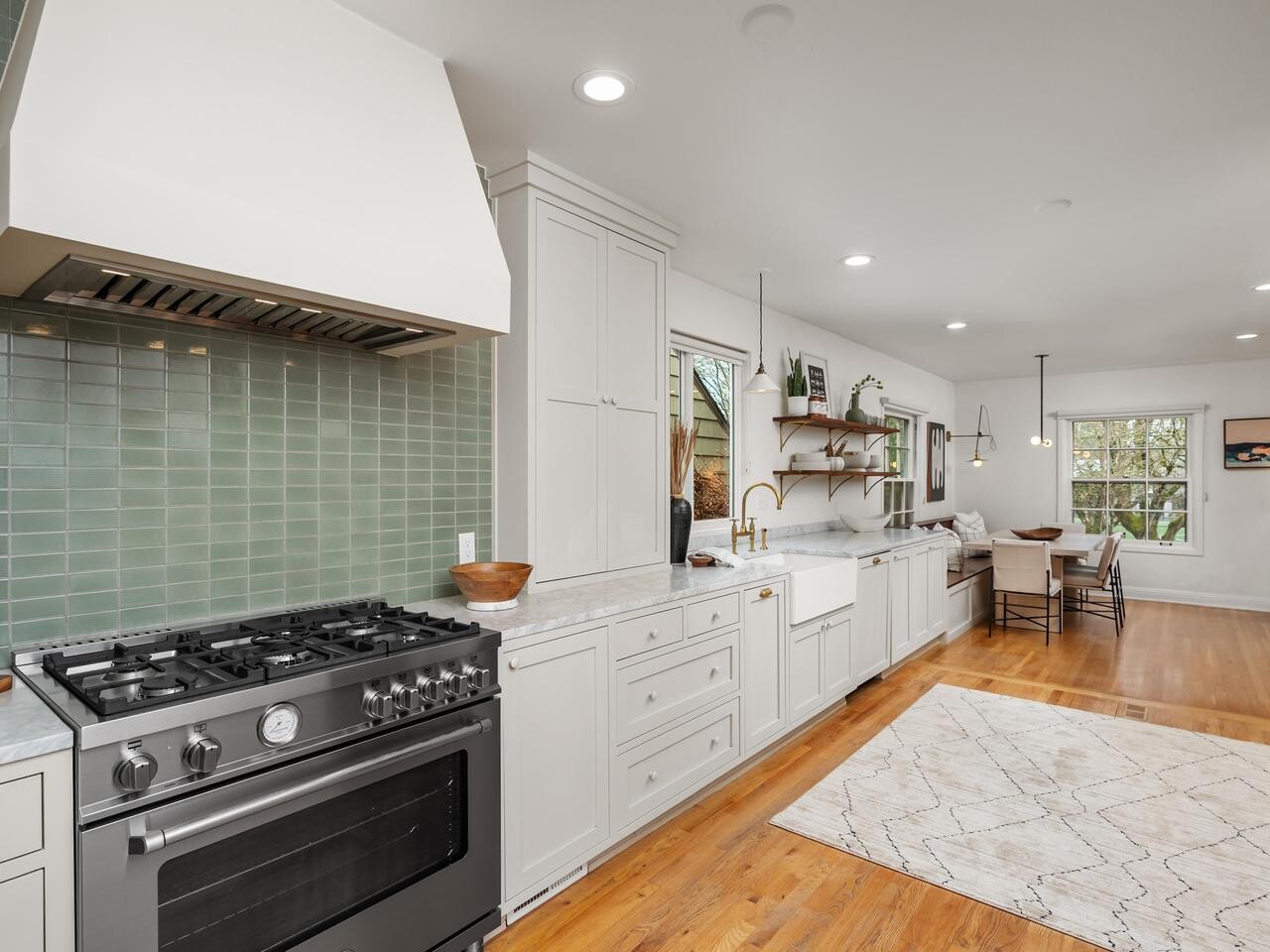 Modern kitchen with light cabinetry, stainless steel gas stove, green tile backsplash, farmhouse sink, open shelves, wooden floor, and a dining area with large windows in the background.
