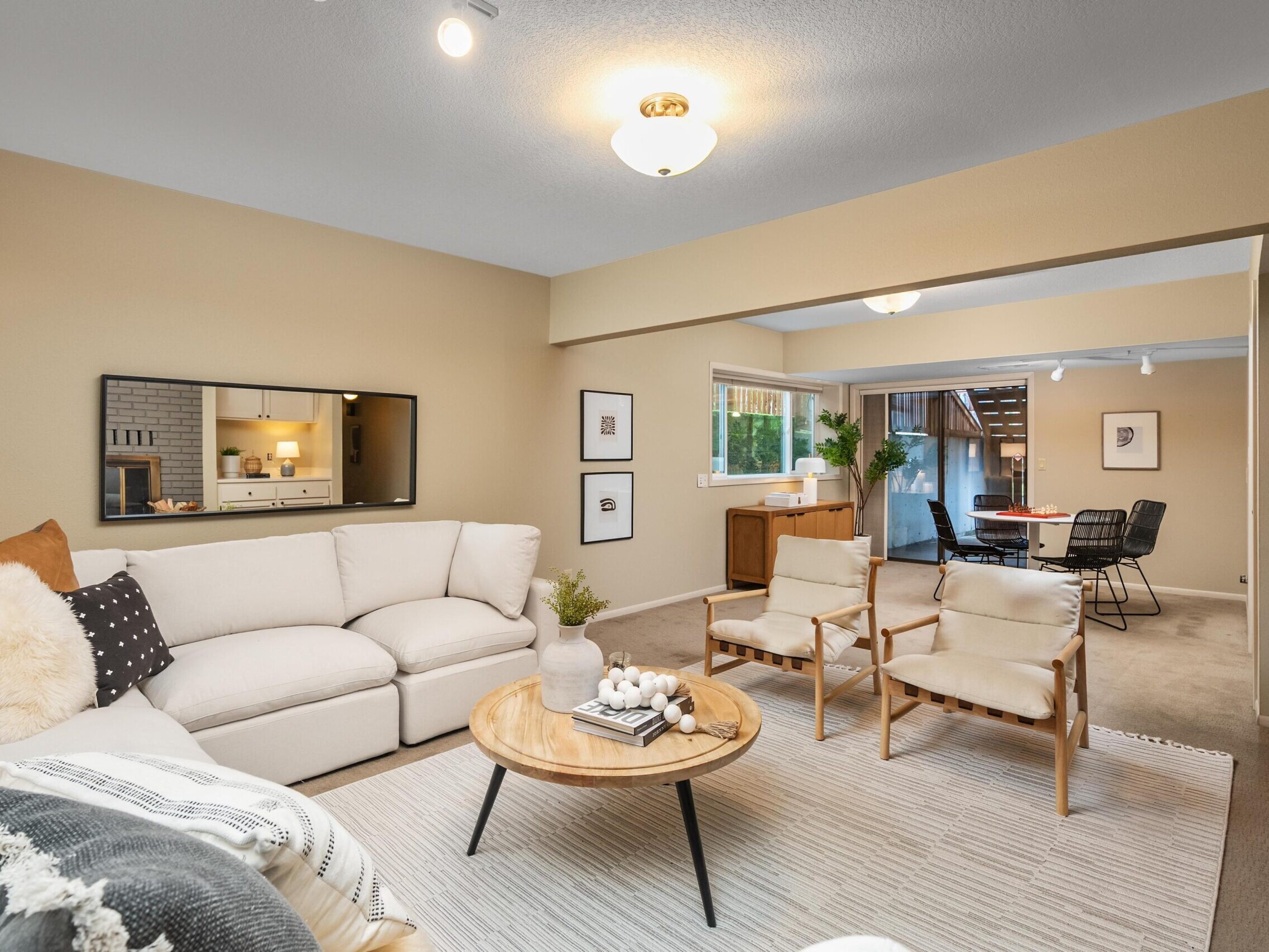 A cozy living room with a white sectional sofa, two beige chairs, a round wooden coffee table, and wall art. The room has neutral tones and leads to a dining area with a table and chairs by large windows.