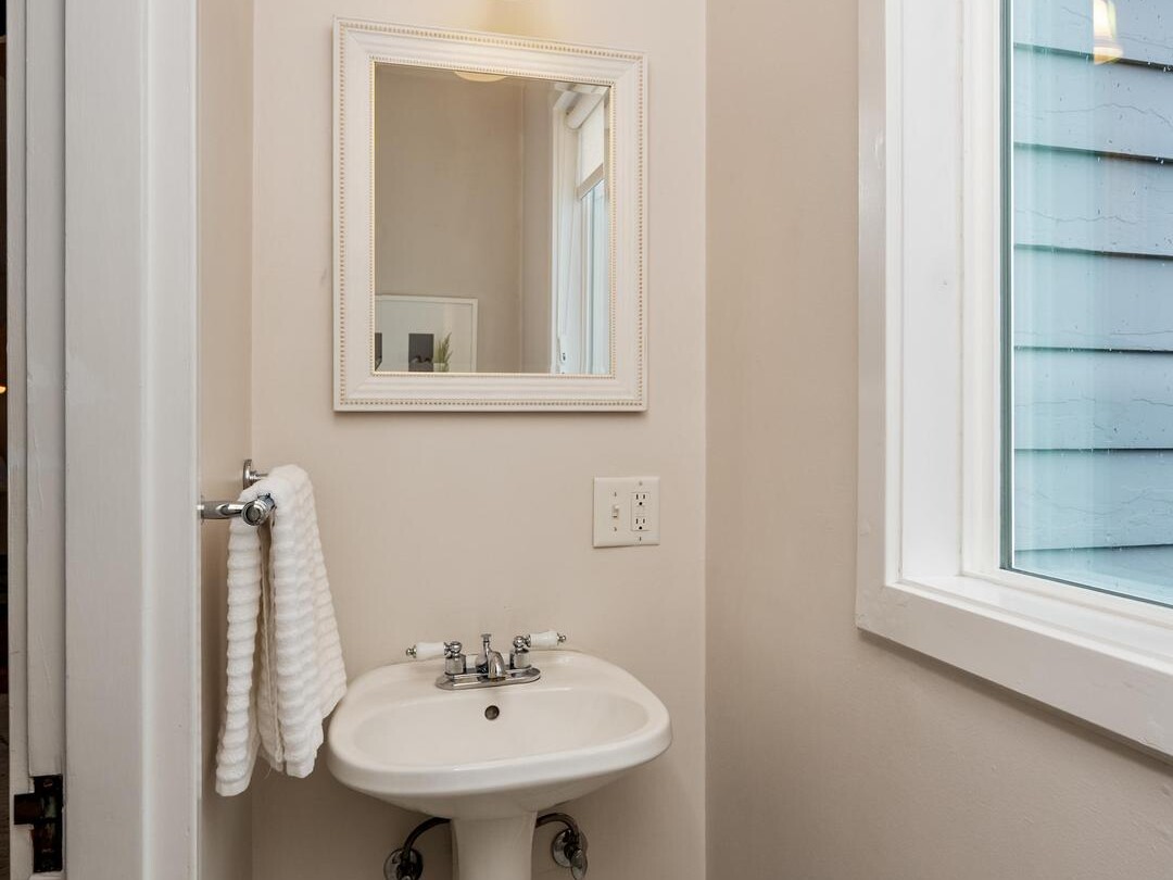 A small bathroom features a white pedestal sink, a wall-mounted mirror with a white frame, a sconce light above, a white towel on a ring, and a large window letting in natural light. The floor has hexagonal tiles.
