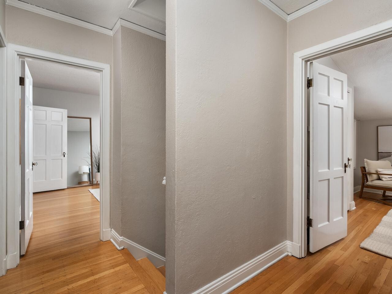 A hallway with light beige walls and wooden floors leads to two rooms with white doors. One room has a chair and a rug; the other features a bed, a plant, and a mirror. The space is clean and well-lit.