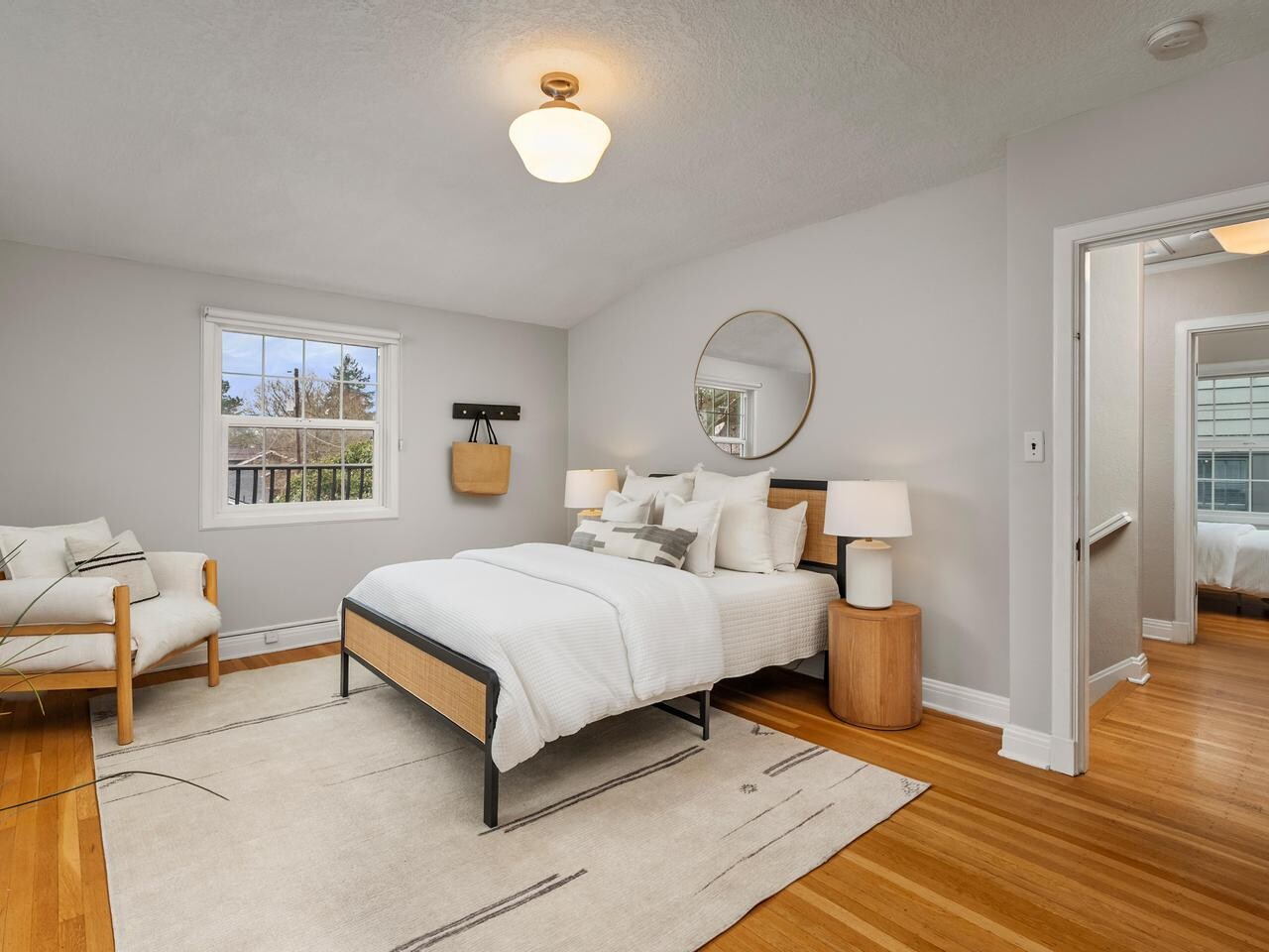 A bright, modern bedroom with light gray walls, wooden floors, a cozy bed with white bedding, two bedside tables with lamps, a round mirror above the bed, and a window letting in natural light.