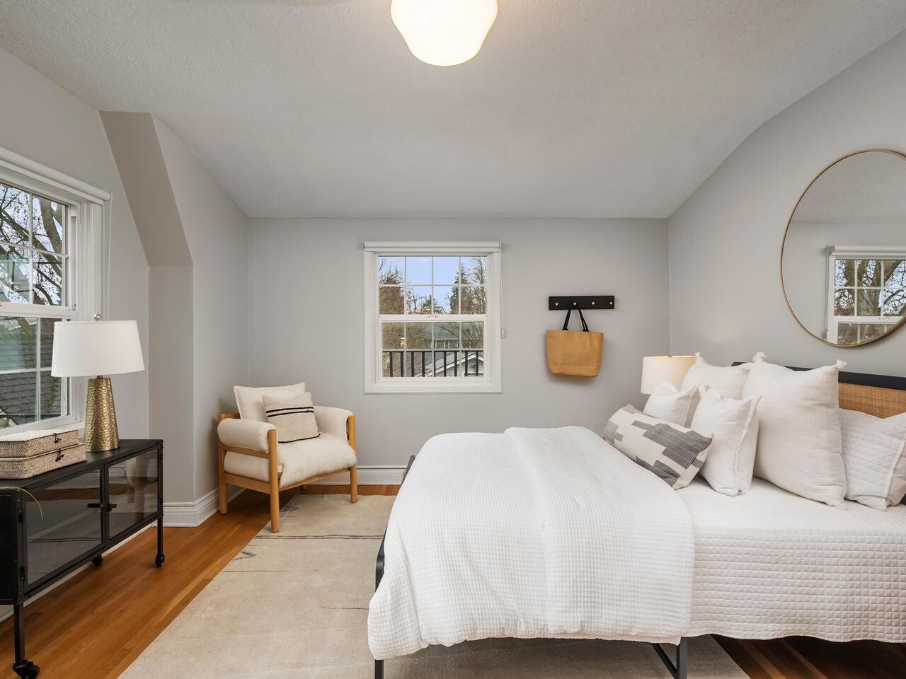 A cozy, modern bedroom with a neatly made bed, white bedding, and several pillows. Theres a round mirror above the bed, a light wood armchair by a window, and a black nightstand with a lamp and books on it.