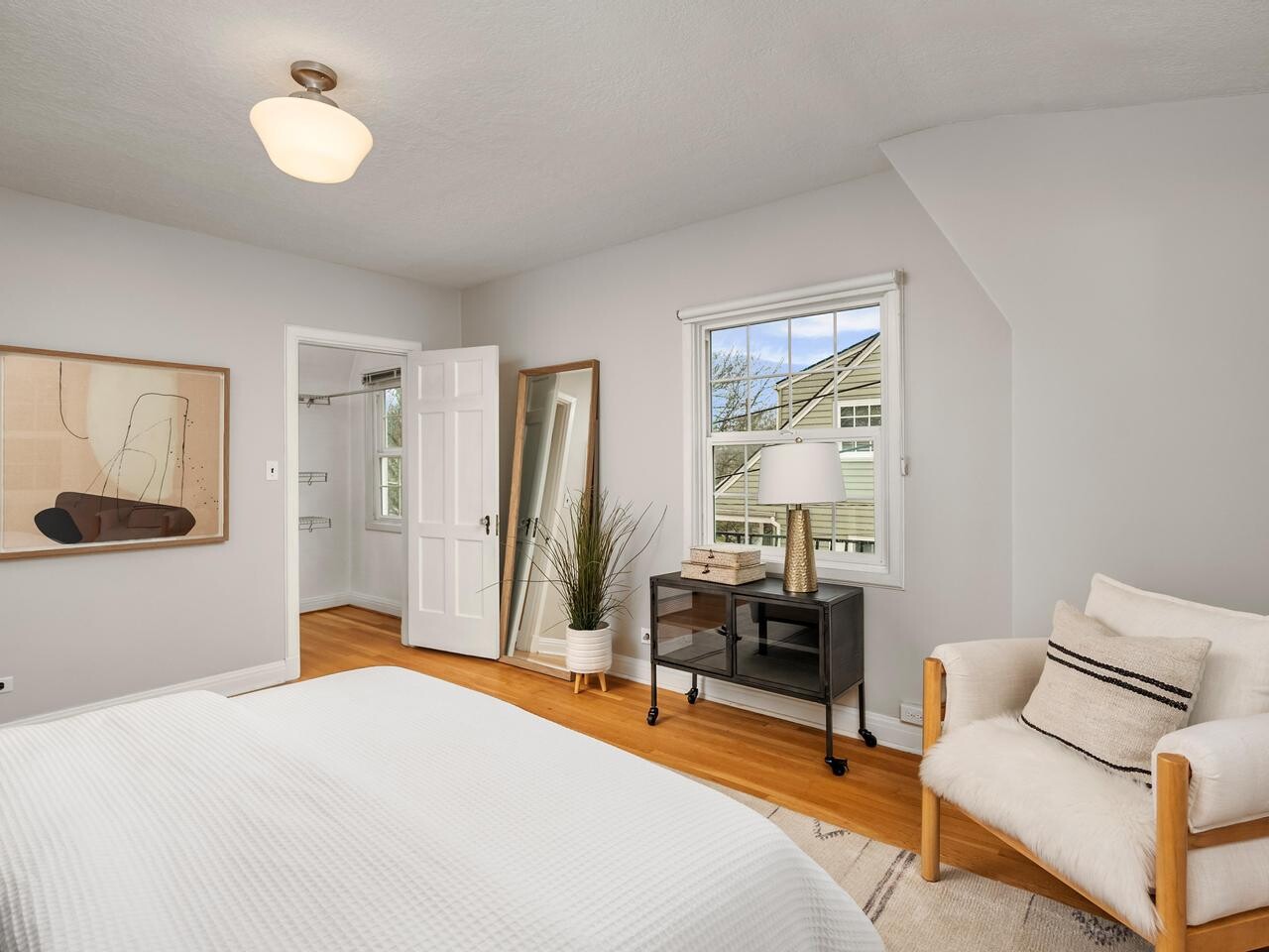 A bright bedroom with light walls, a bed with a white blanket, an armchair with pillows, a black dresser with a lamp, a potted plant, abstract wall art, and a window letting in daylight.