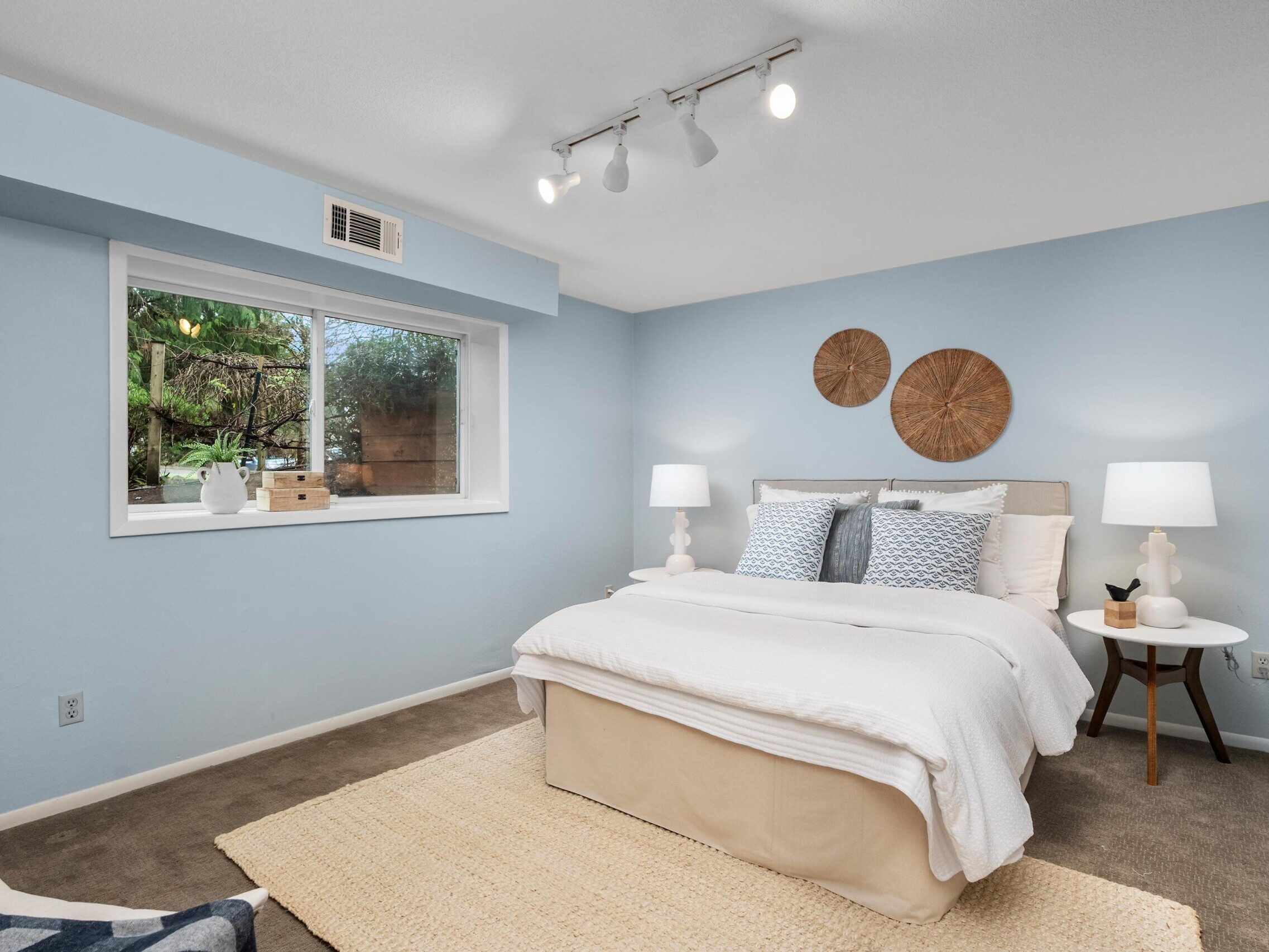 A cozy bedroom with light blue walls, a large window, a white bed with blue and white pillows, two white lamps on bedside tables, a beige rug, and decorative wall art above the bed.