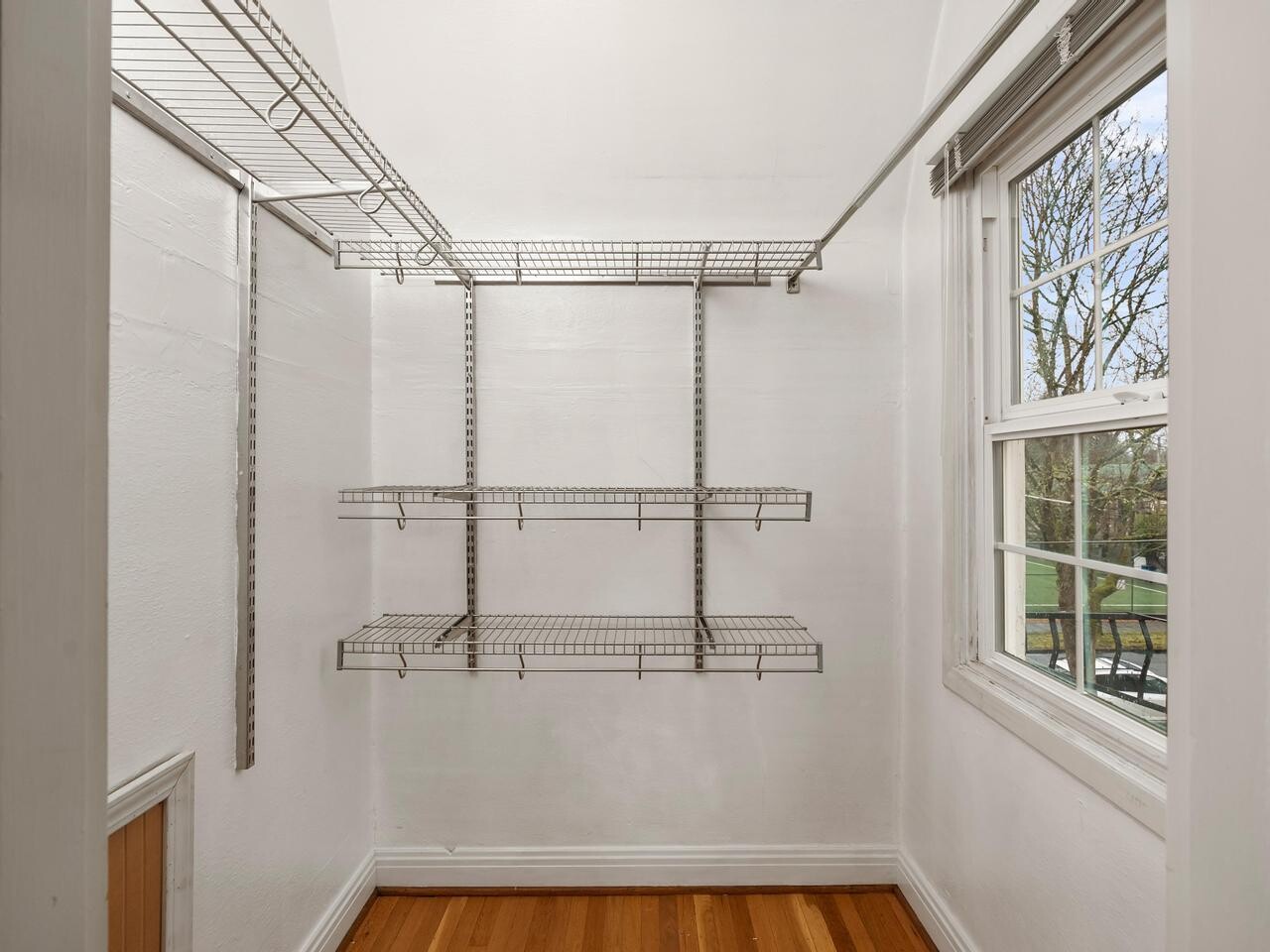 A small walk-in closet with white walls, metal wire shelving on the left and back walls, a hardwood floor, and a window on the right letting in natural light.