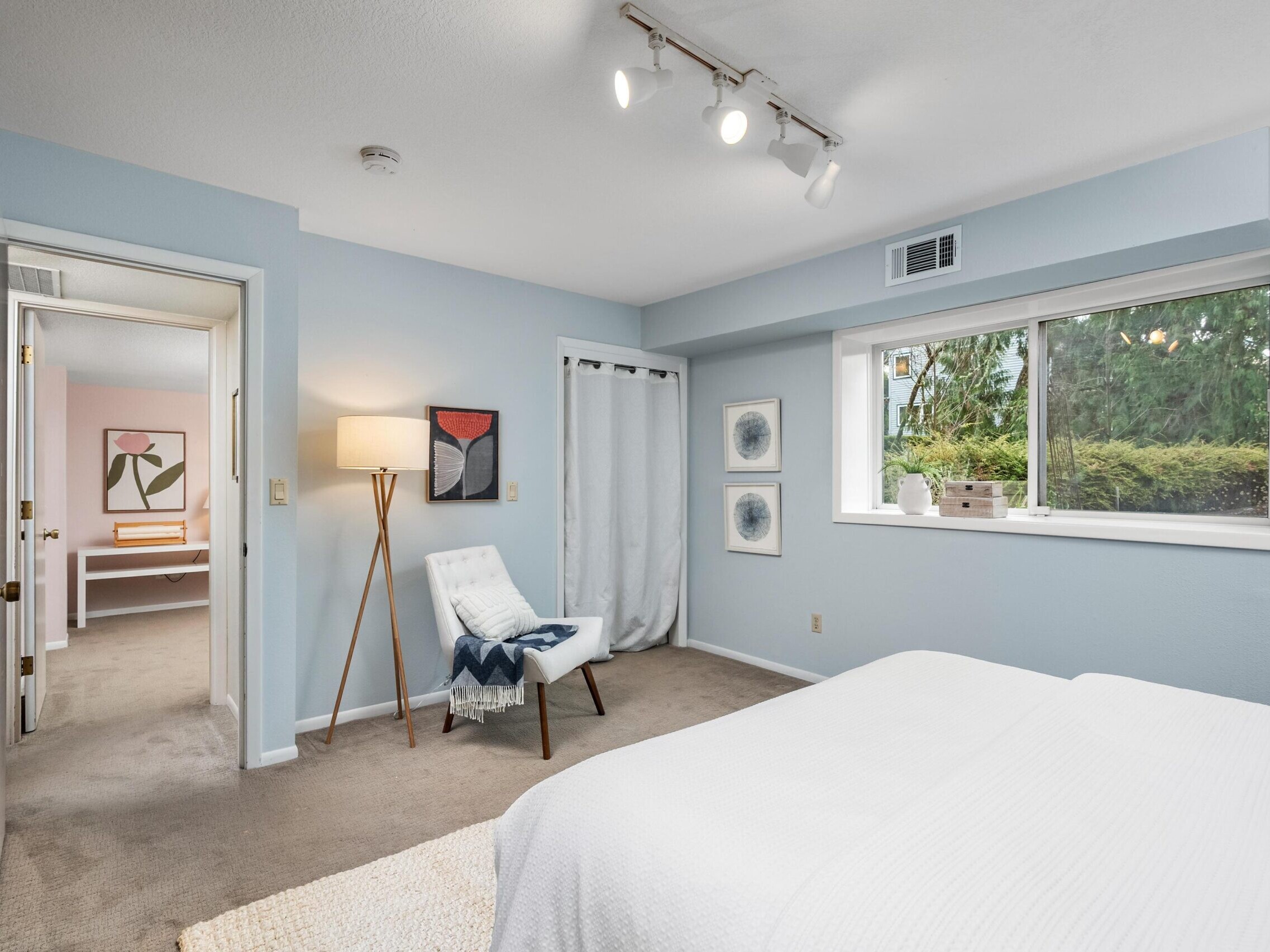 A bright bedroom with light blue walls, a bed with white bedding, a white chair with a blue pillow, a floor lamp, wall art, a window with greenery outside, and an open door leading to another room.