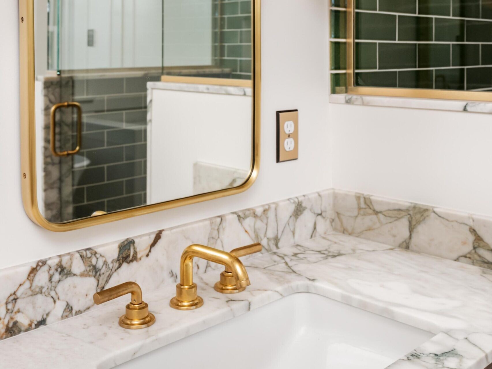 A bathroom vanity with a white and gray marble countertop, gold faucet and fixtures, a rectangular gold-framed mirror, green subway tile backsplash, and a glass shower enclosure reflected in the mirror.