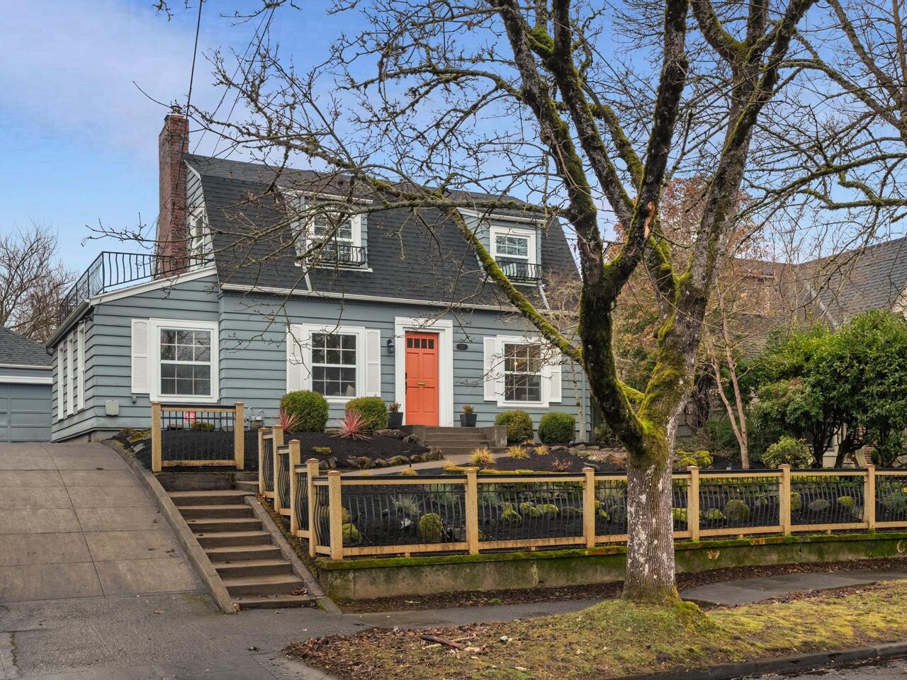 A two-story blue-gray house with a dark roof, white trim, and a coral front door sits behind a wooden fence. A bare tree stands in the front yard, and a driveway slopes up on the left side.