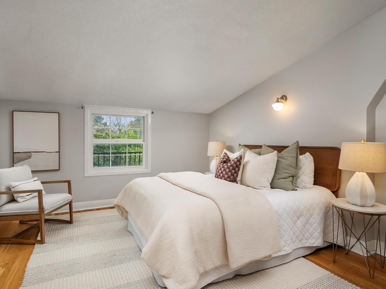 A cozy bedroom with a large bed, multiple pillows, two bedside lamps, a cushioned chair, a framed artwork, a window with a view of greenery, light walls, and a striped rug on a wooden floor.