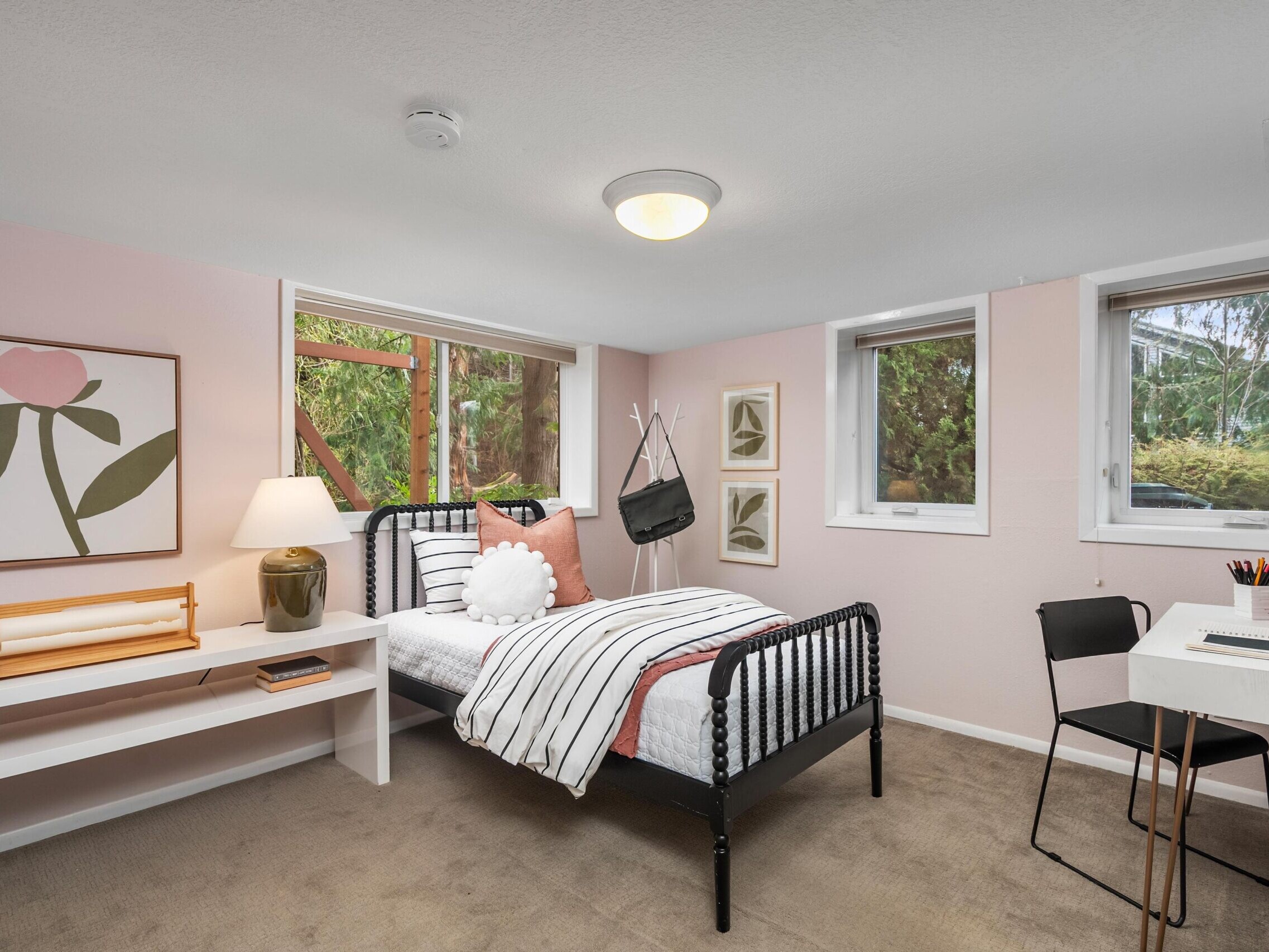 A cozy bedroom with light pink walls, a single black metal bed with striped bedding, a desk and chair, wall art, a lamp, and large windows showing greenery outside.