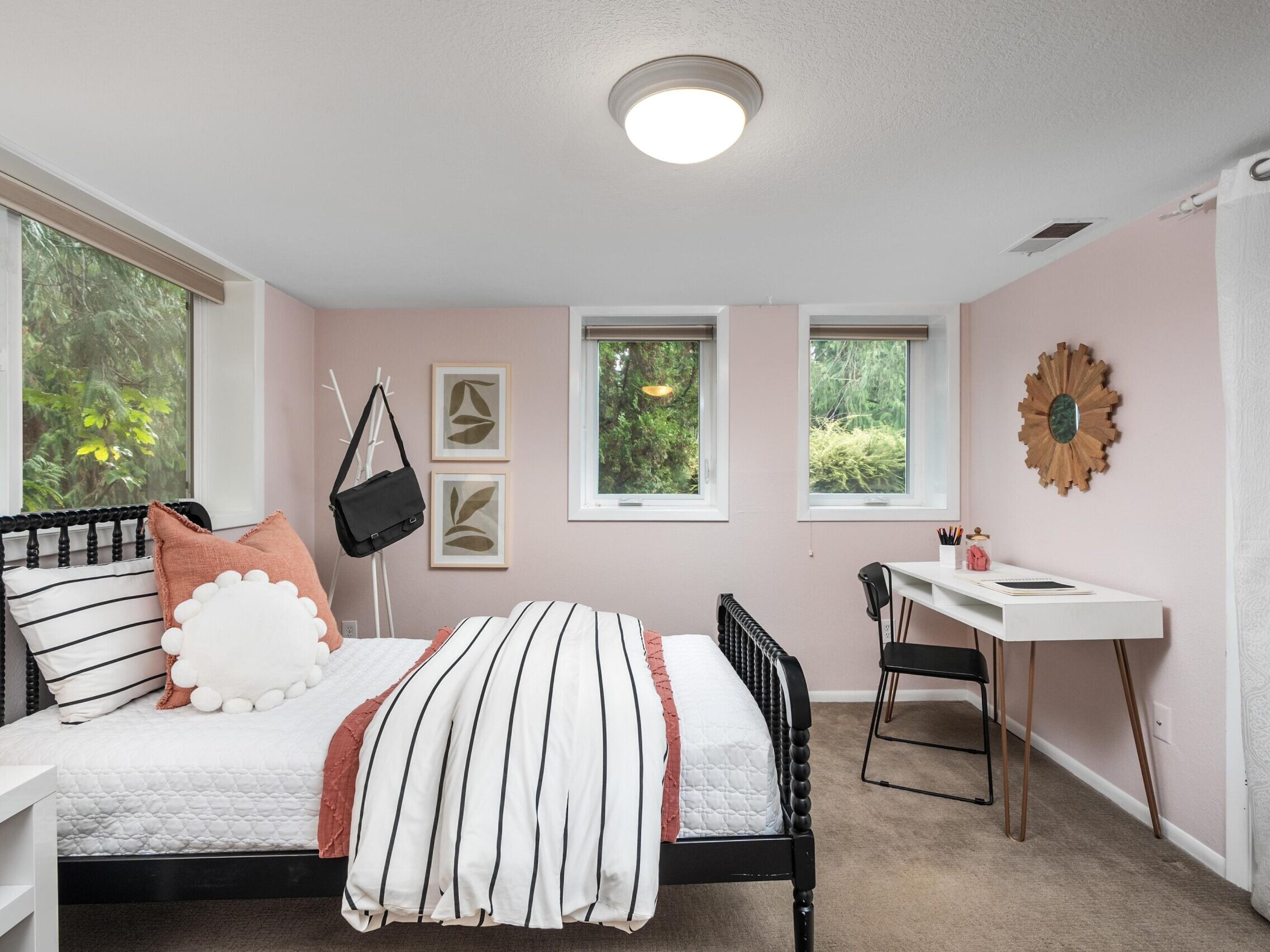 A cozy bedroom with pale pink walls, featuring a black metal bed with striped bedding, decorative pillows, a small desk with chair, two windows, framed art, and a round sunburst mirror on the wall.