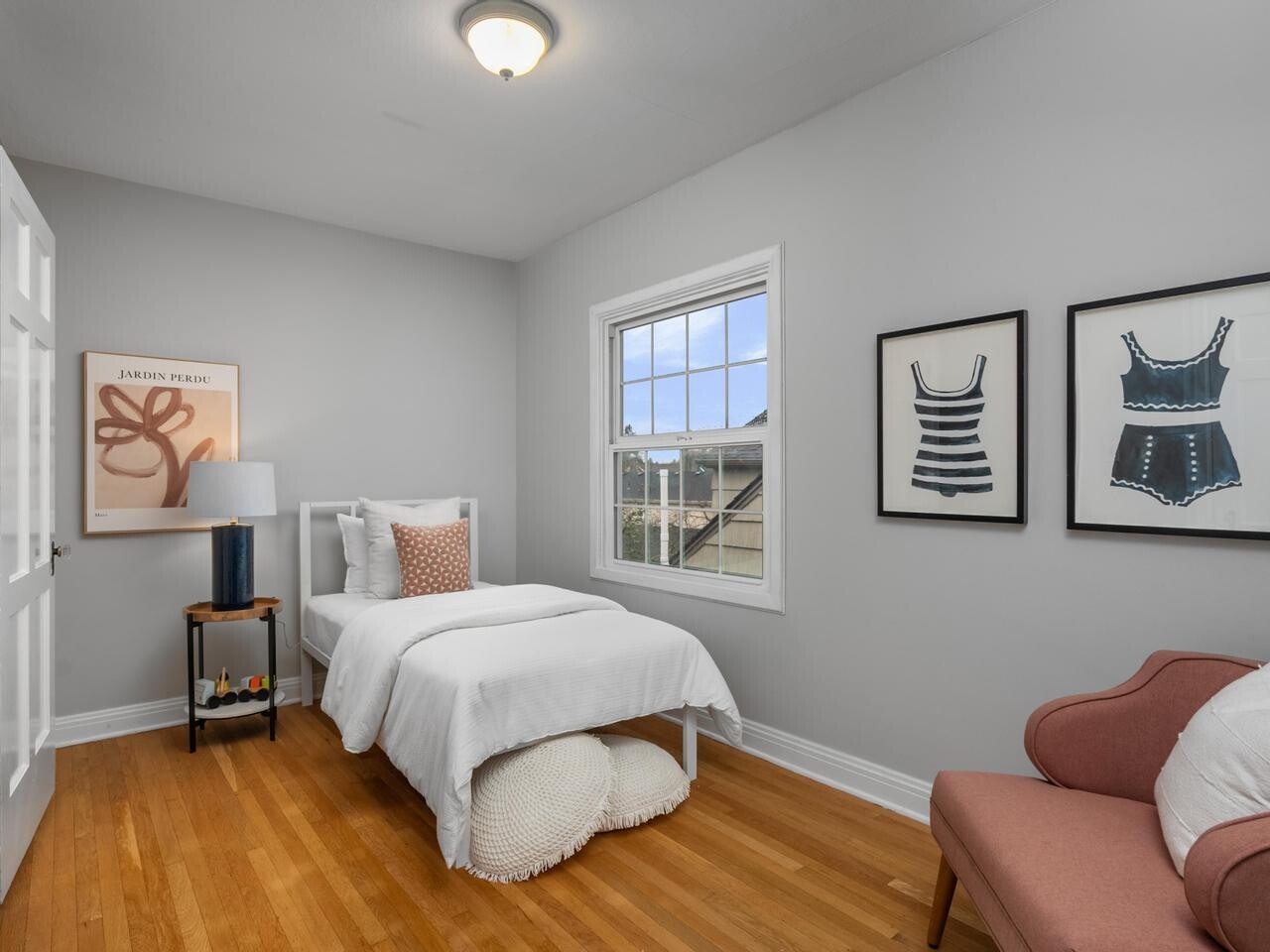 A small, neatly arranged bedroom with a single bed, white bedding, a side table with a lamp, a pink armchair, and framed swimsuit artwork on gray walls. A window lets in natural light.