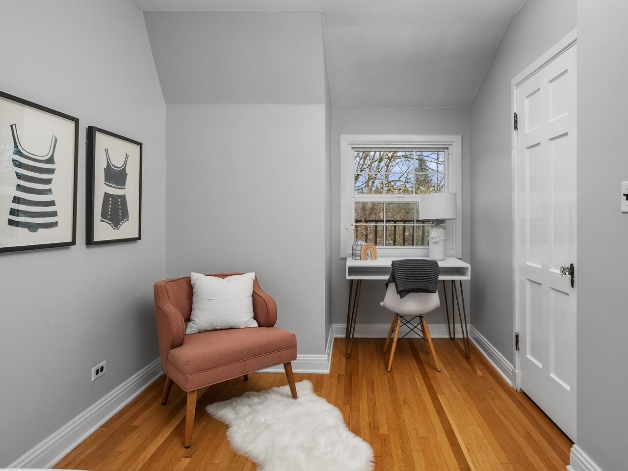 A small home office with a white desk and chair by a window, a peach armchair with a white rug, and two framed swimsuit prints on the light gray wall. The room has hardwood floors and a closed white door.