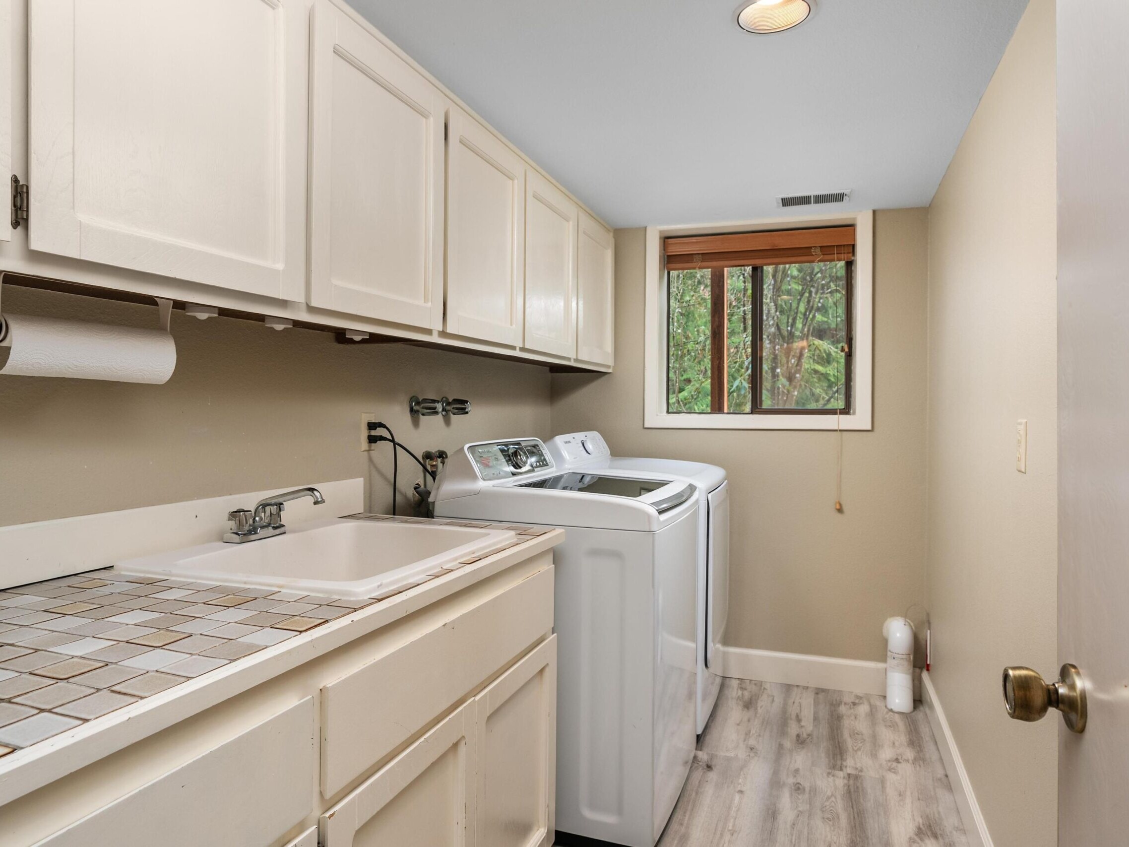 Laundry room with white cabinets, a tiled countertop with a sink, a paper towel holder, a washer and dryer, light wood floors, beige walls, and a window with a view of trees.