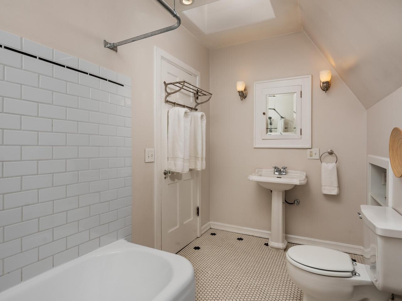 A clean, white bathroom with a bathtub, pedestal sink, toilet, wall-mounted mirror, towel racks with white towels, hexagonal floor tiles, and two wall sconces; natural light from a skylight above.