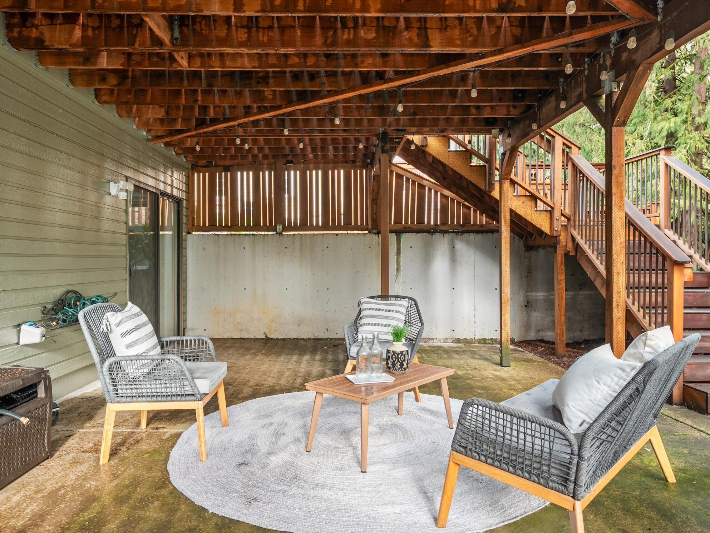 A cozy covered patio features two cushioned chairs, a loveseat, and a round rug around a wooden coffee table. String lights hang overhead, and wooden stairs lead up to a deck. Greenery is visible in the background.
