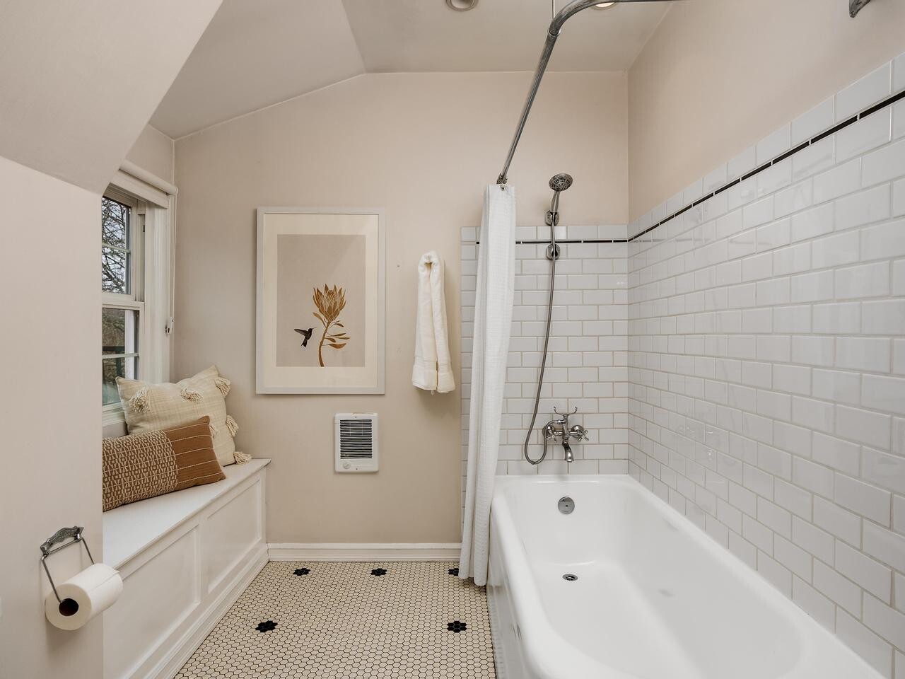 A cozy bathroom with a white bathtub, white subway tile walls, hexagon tile floor, a shower curtain, towel, window seat with cushions, wall art, and a toilet paper holder on the left.