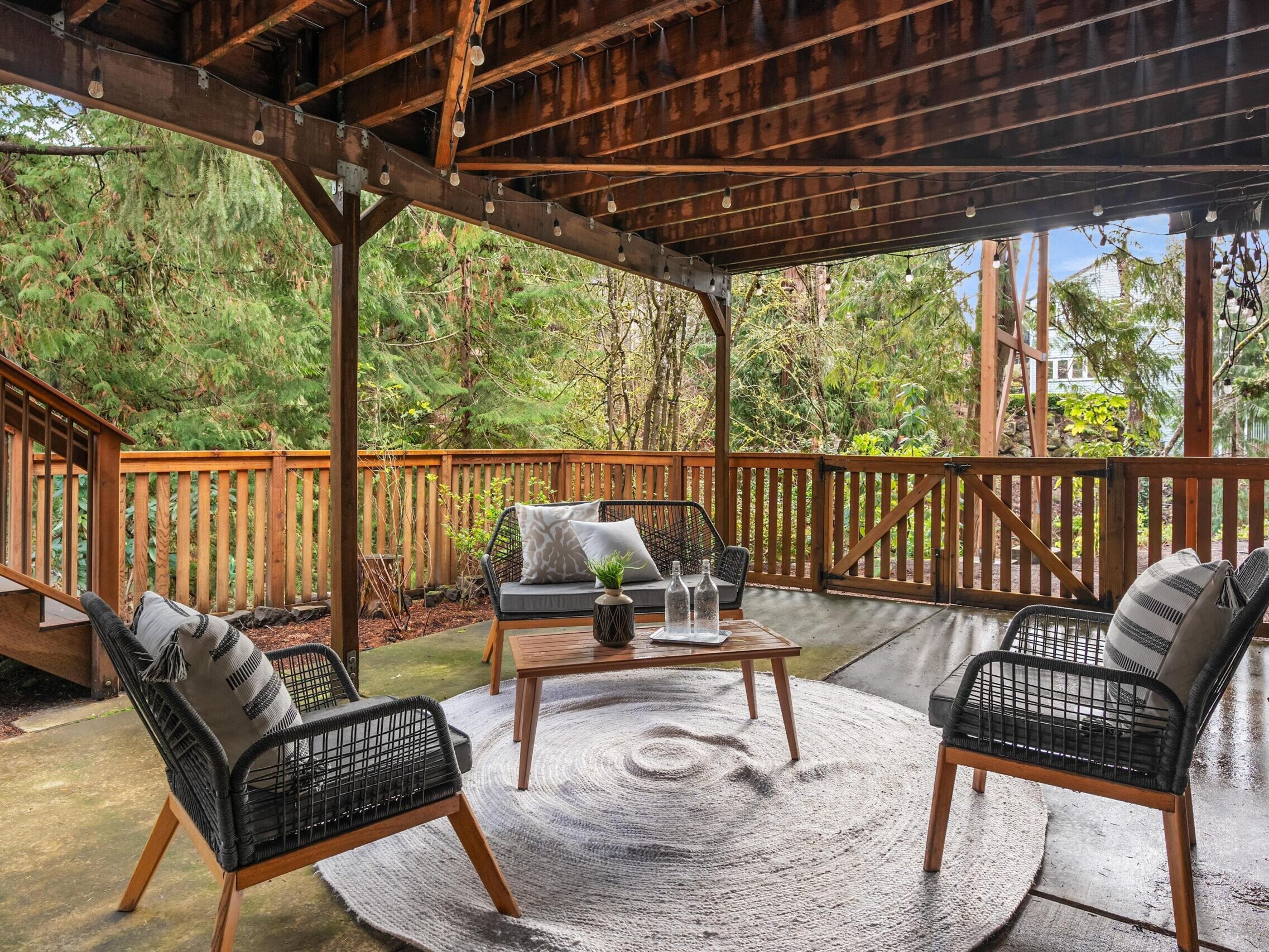 A covered outdoor patio with four black wicker chairs and a small table on a round rug. Wooden railings and stairs lead to a wooded backyard. String lights hang from the exposed wooden ceiling.