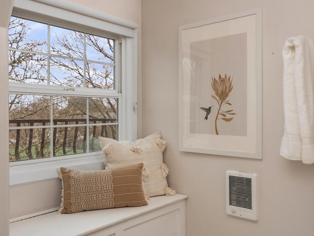 A cozy window seat with beige and cream pillows sits next to a large window in a light-colored bathroom. A towel hangs on the wall and a botanical art print is displayed above a small heater on white hex tile flooring.