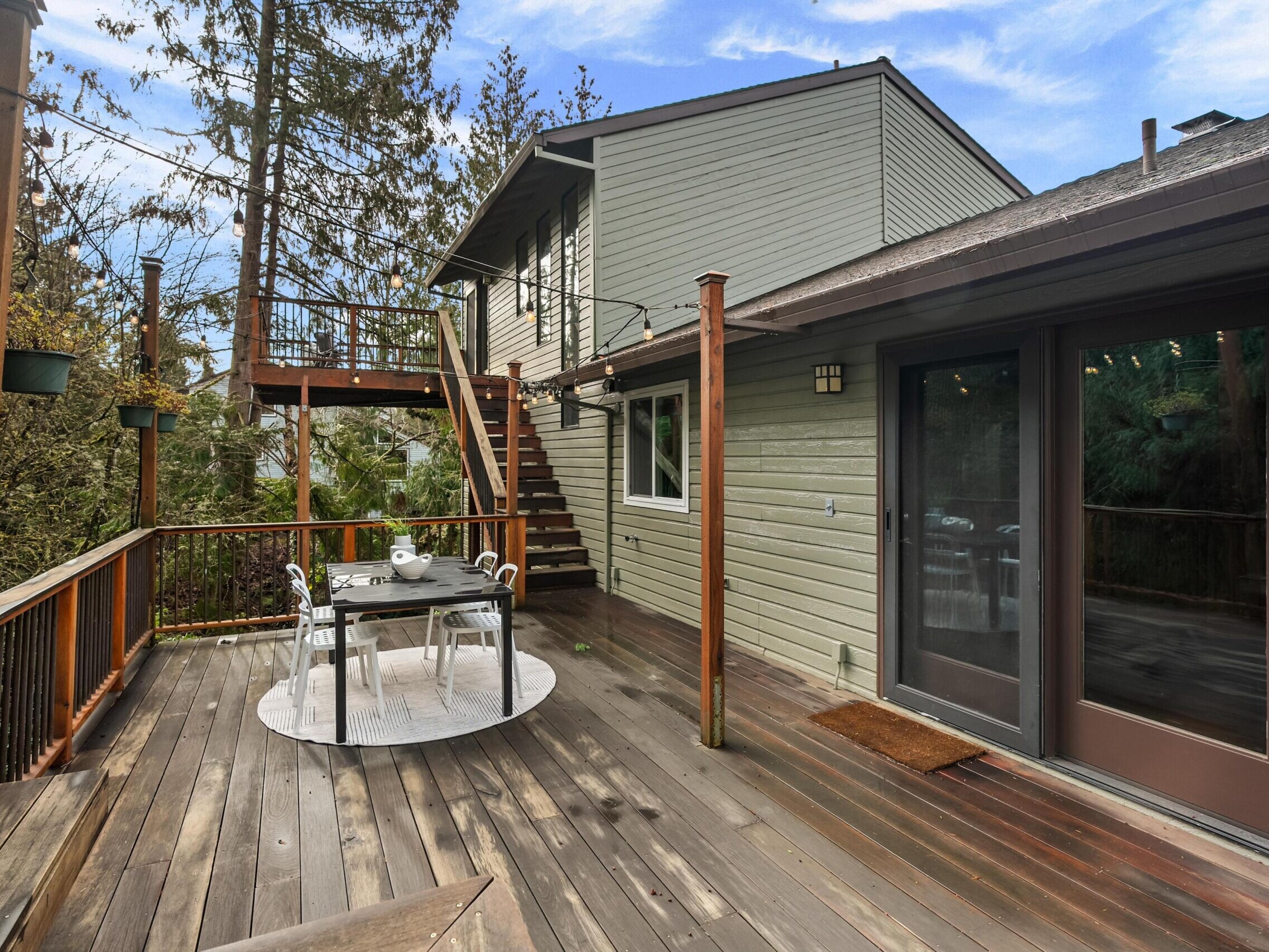 A spacious wooden deck with a dining table and chairs, bordered by railings and surrounded by trees, leads to a staircase ascending to an upper balcony attached to a green two-story house.