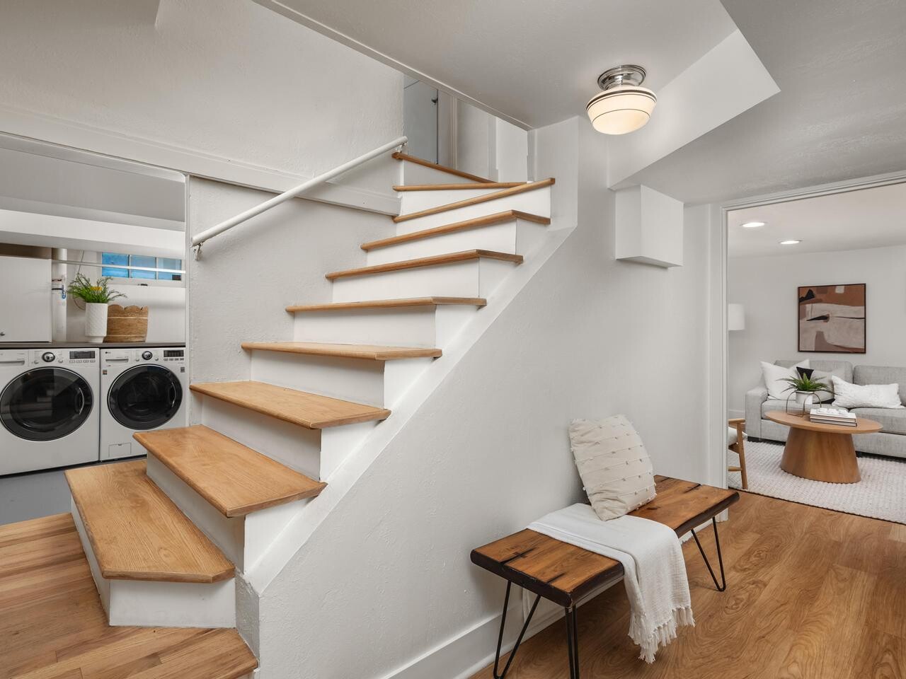 A modern, bright interior featuring a wooden staircase, a bench with a cushion and throw, a laundry area with washer and dryer, and a cozy living room with a sofa and coffee table in the background.