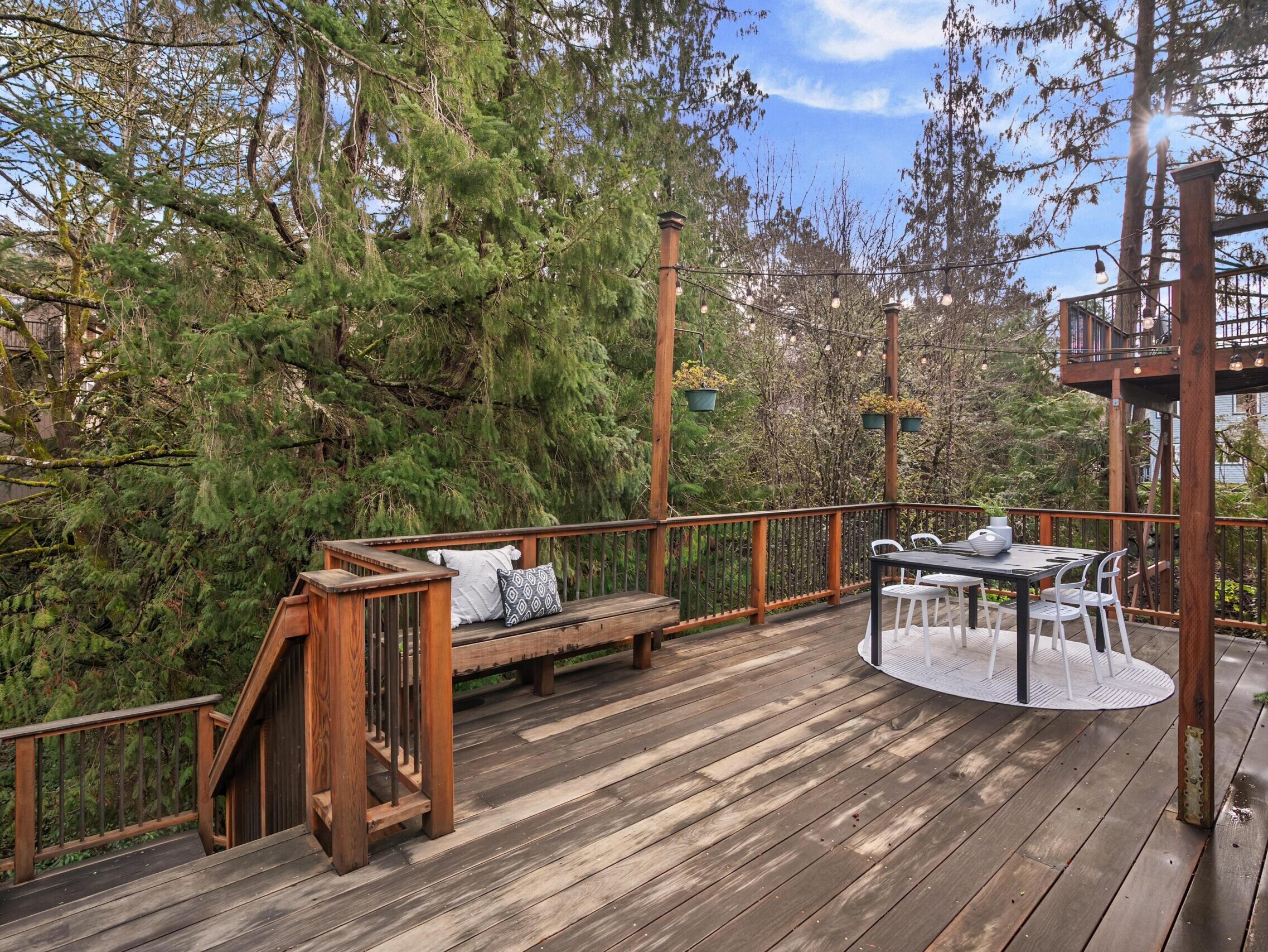 A spacious wooden deck surrounded by tall trees features a bench with cushions, a dining table with chairs on a round rug, and string lights overhead, creating a cozy outdoor seating area.