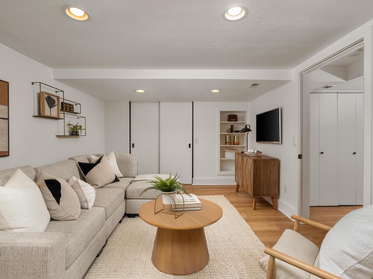 A modern living room with a beige sectional sofa, neutral pillows, a round wooden coffee table, and a light rug. Wall art, a TV on a wooden console, shelves with books, and sliding closet doors are visible.