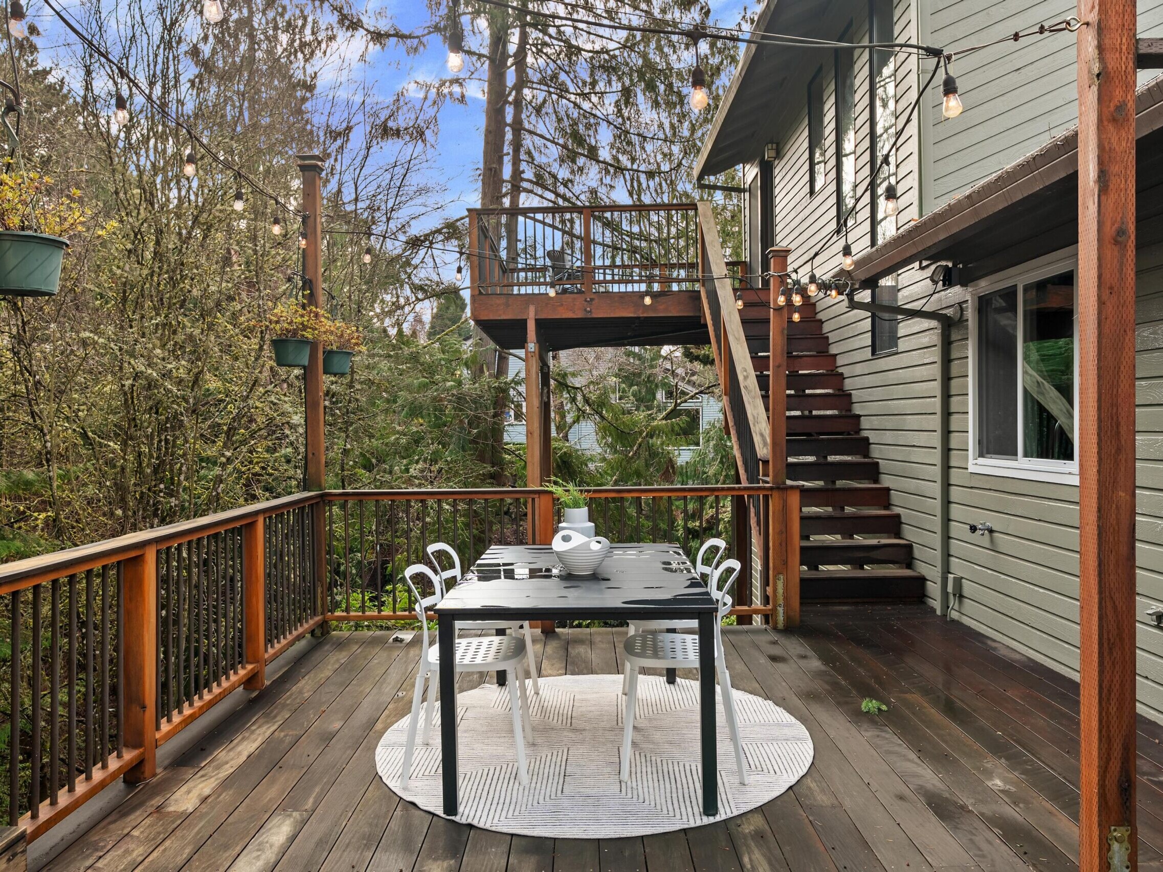 A wooden deck with a round table and four chairs, set with dishes, surrounded by trees. String lights hang above, and stairs lead to an upper balcony attached to a gray house.
