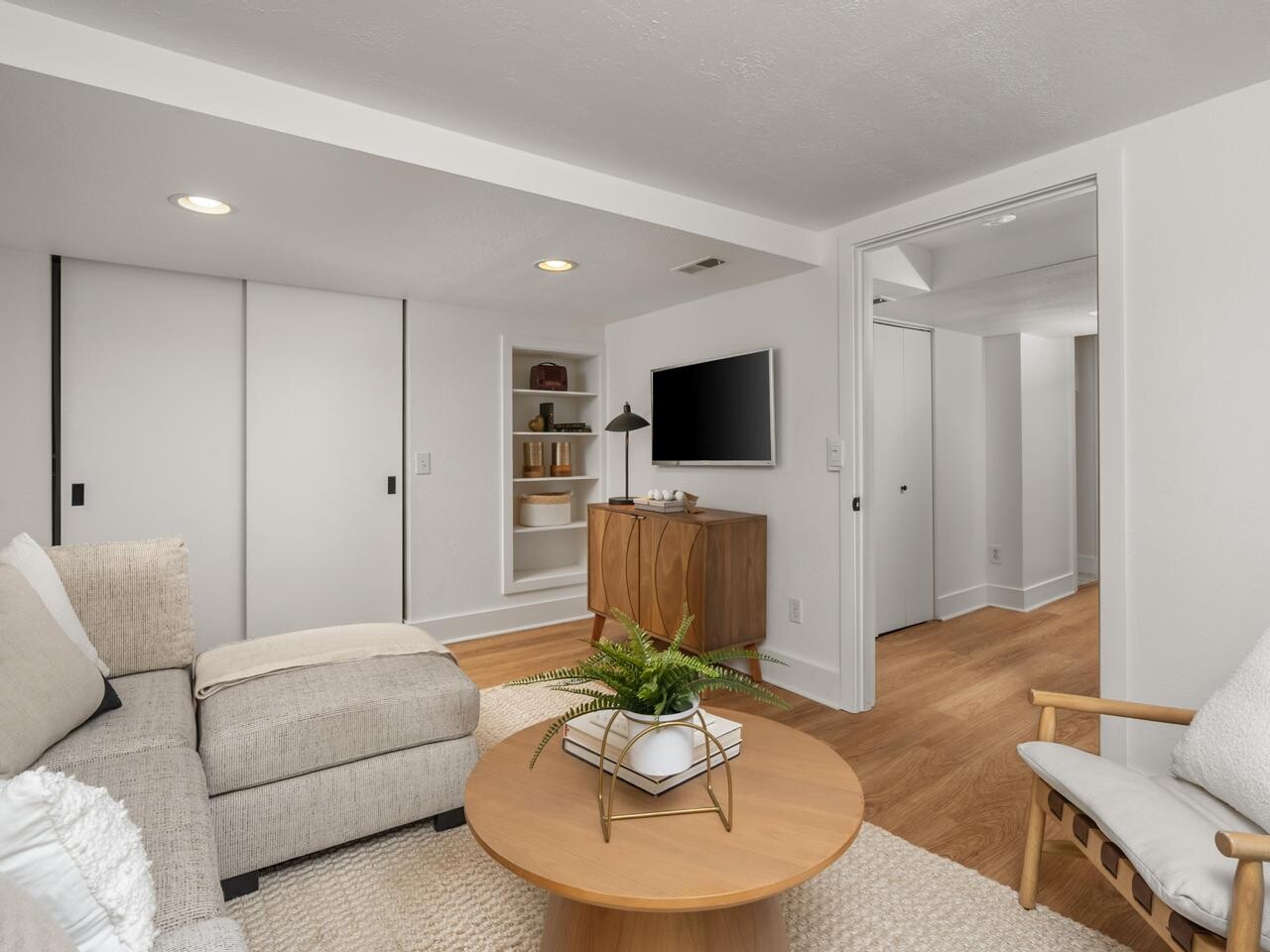 A cozy, modern living room with a beige sectional sofa, a round wooden coffee table with a plant, a flat-screen TV on a wooden console, built-in shelves, and light wood flooring. The room features white walls and sliding closet doors.