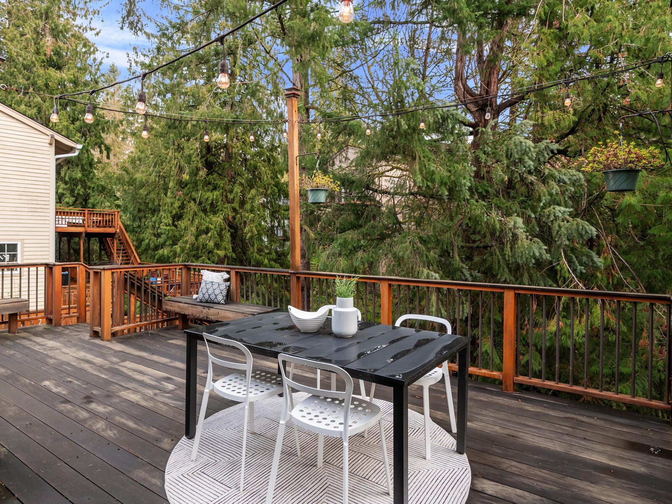 A wooden deck with a black table, four white chairs, a decorative centerpiece, string lights overhead, and surrounded by trees and greenery. A bench sits in the background near a house.
