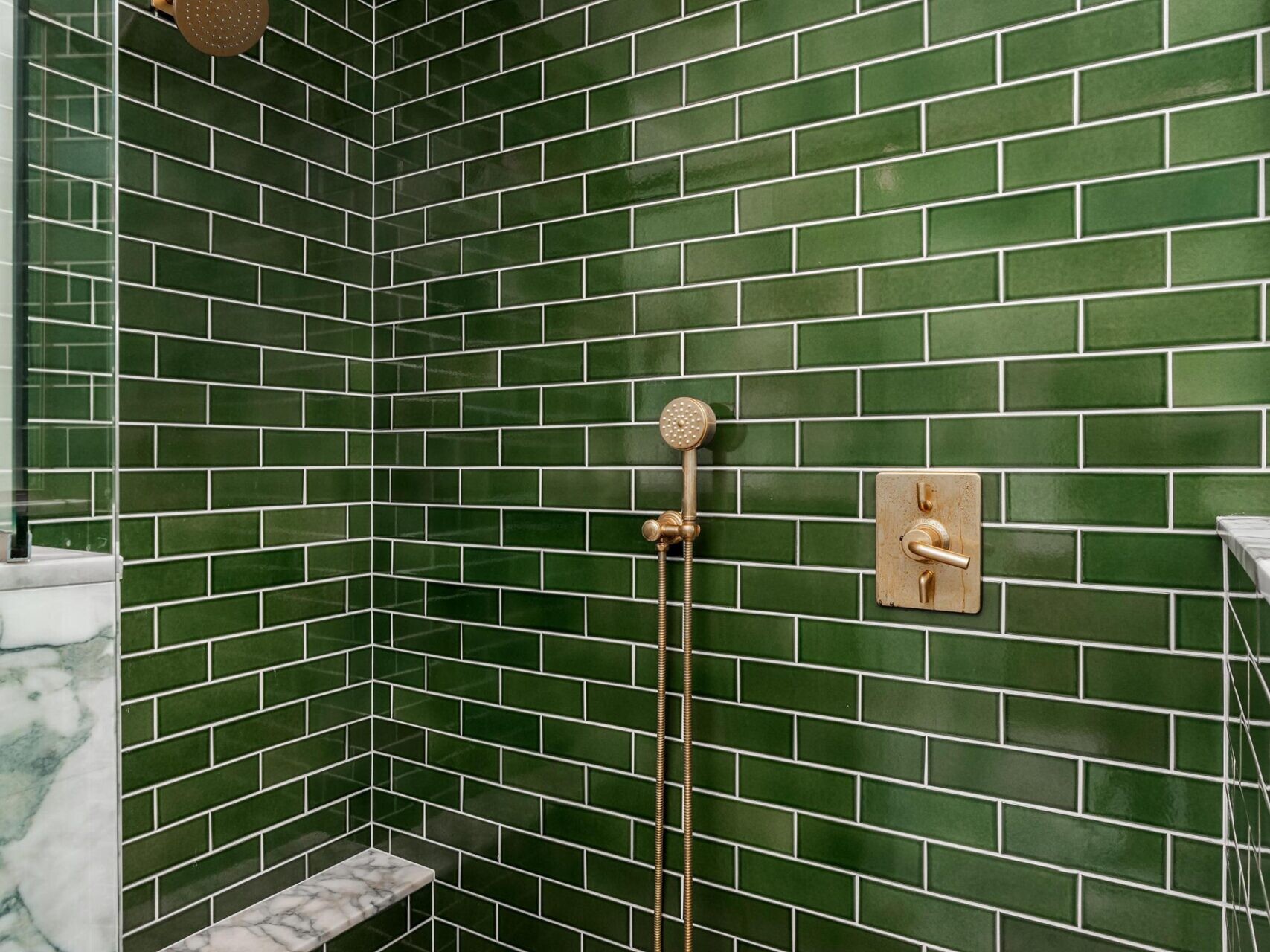 A modern shower with glossy green subway tiles, a marble bench, and gold fixtures including a rainfall showerhead, handheld shower, and faucet; the floor has a light herringbone tile pattern.