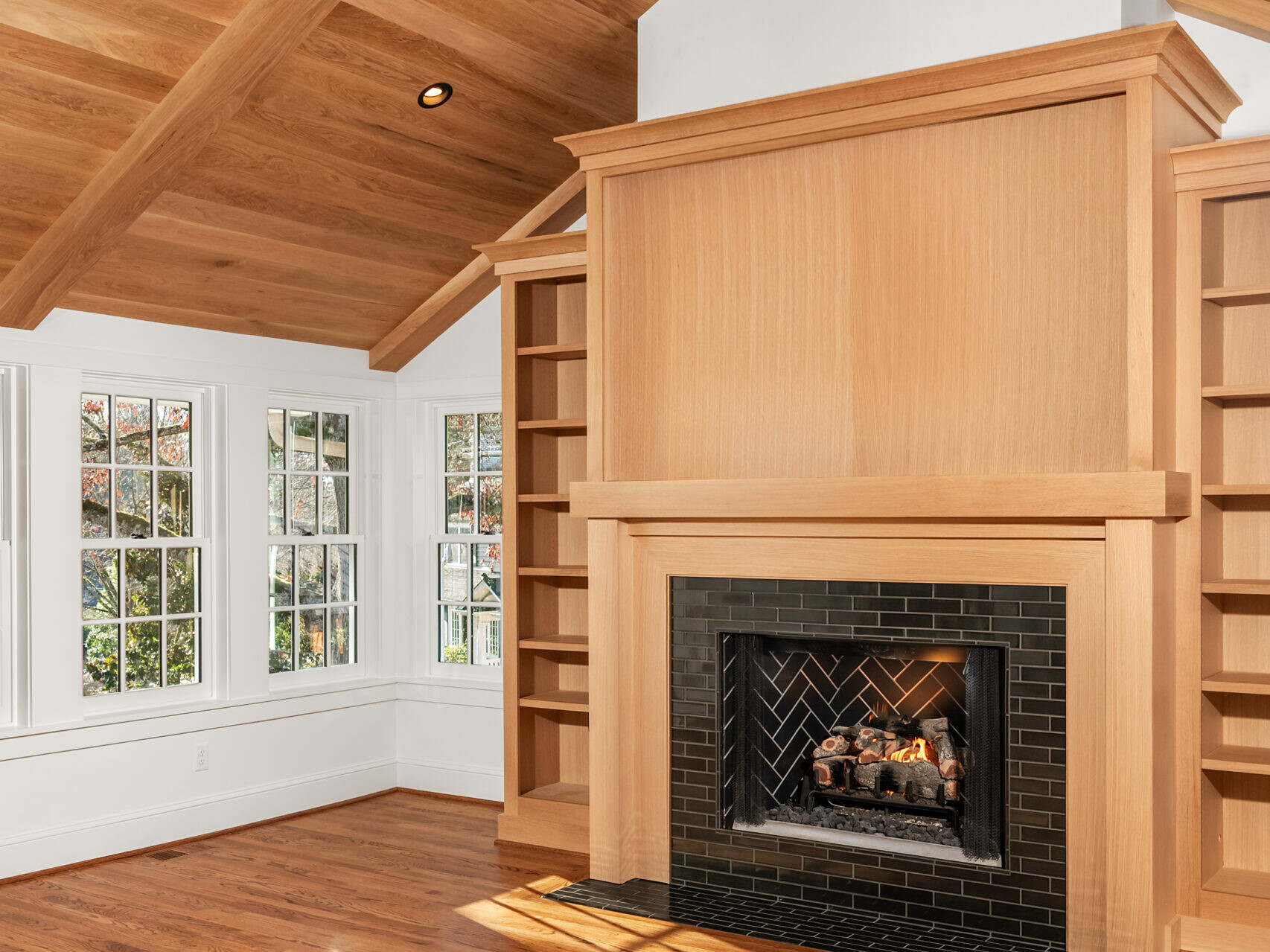 A bright, modern living room with a wooden vaulted ceiling, large windows, built-in shelves, and a fireplace with a black tile surround. Sunlight casts shadows on the hardwood floor.