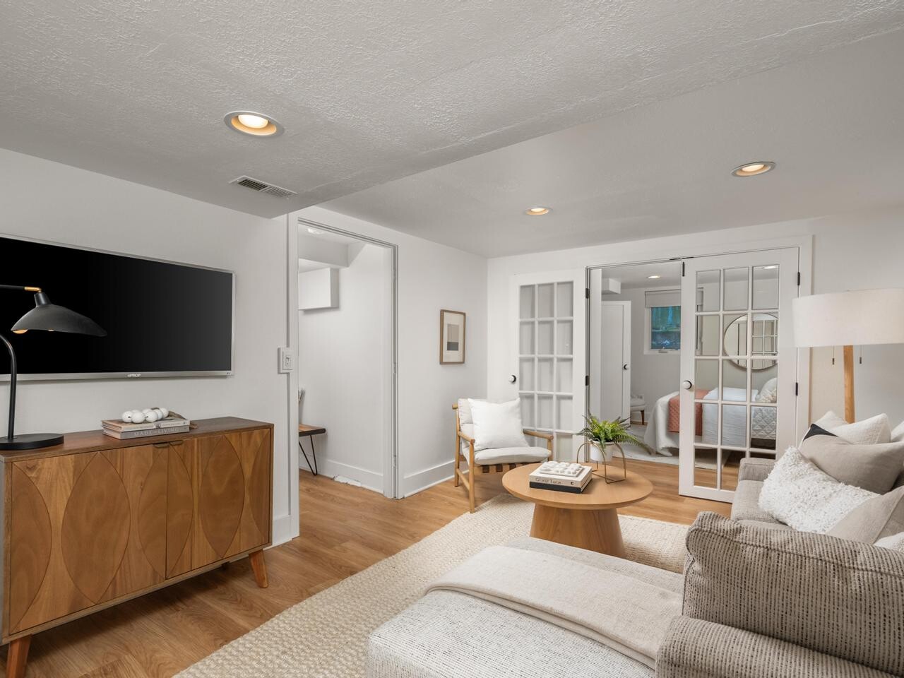 Modern living room with a wall-mounted TV, wooden console, neutral sofa, round coffee table, armchair, and floor lamp. French doors lead to a bright bedroom in the background. Light walls and wood floors create an airy feel.
