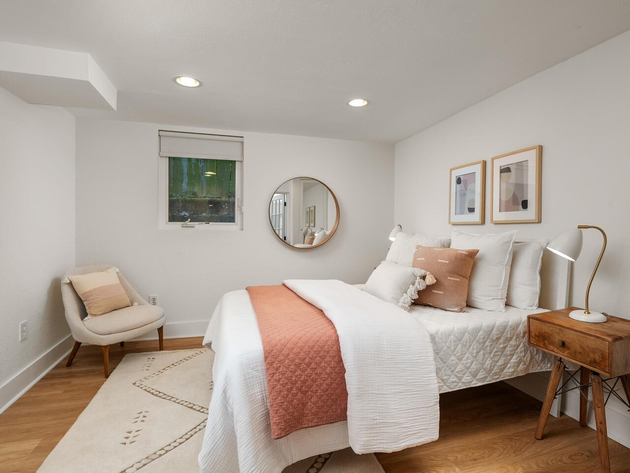 A cozy bedroom with a bed featuring white and blush bedding, a wooden side table with a lamp, a modern chair with pillows, wall art, a round mirror, and a window with a white shade. The room has light walls and wood flooring.