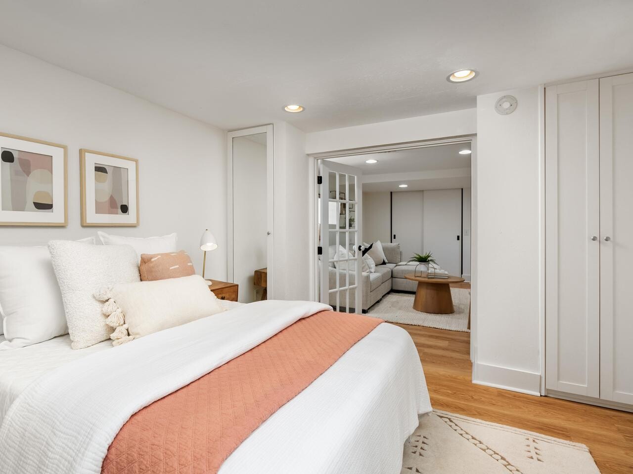 A modern bedroom with white walls, a bed with white and peach bedding, abstract art above the headboard, wood floors, and a view into a cozy sitting area through open French doors.