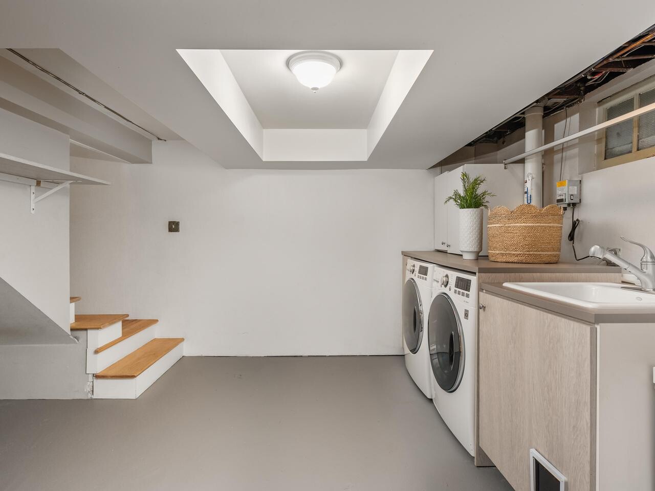 A clean, bright basement laundry room with a washer, dryer, utility sink, light fixture, staircase, open shelving, and a potted plant on the counter. The walls and floor are painted in neutral tones.