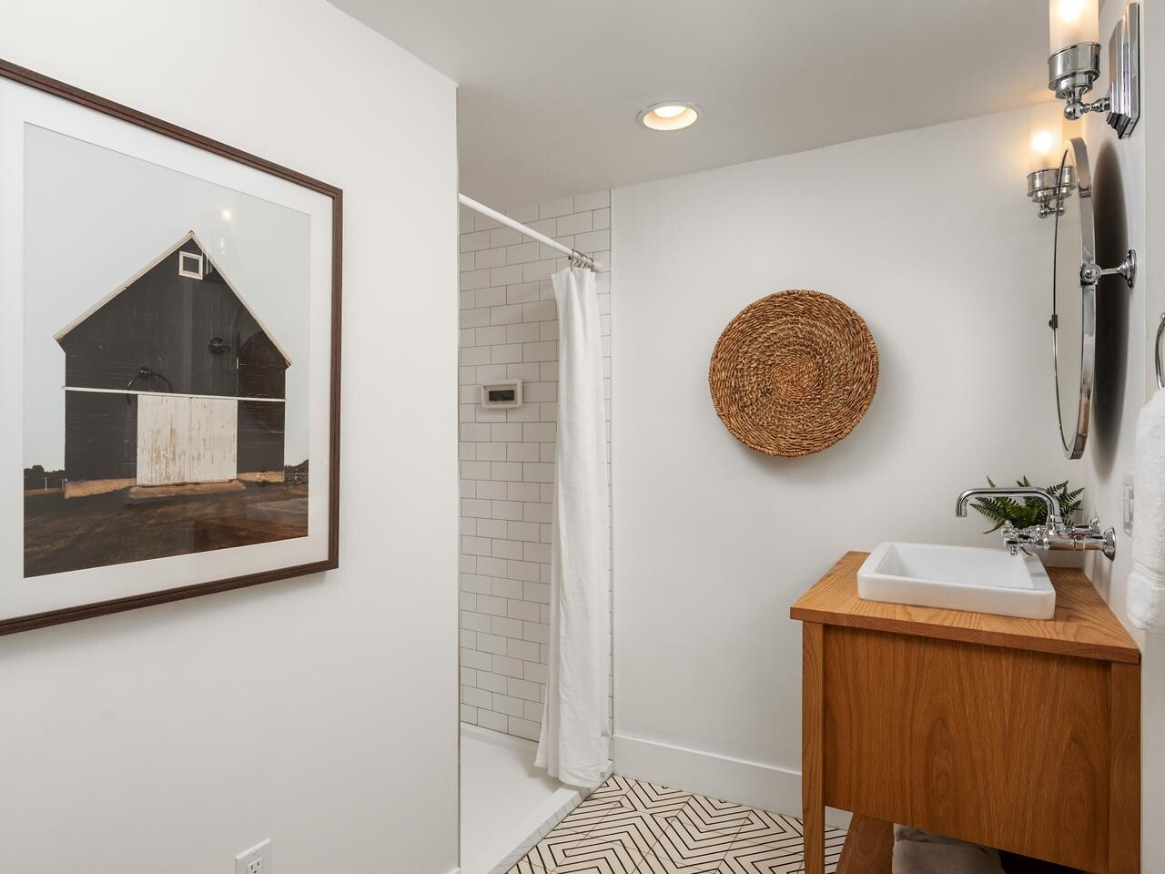 A modern bathroom with white walls, a wooden vanity with a vessel sink, a round mirror, wall-mounted lights, a woven wall decor, and a walk-in shower with white subway tiles. A framed barn photo hangs on the left wall.
