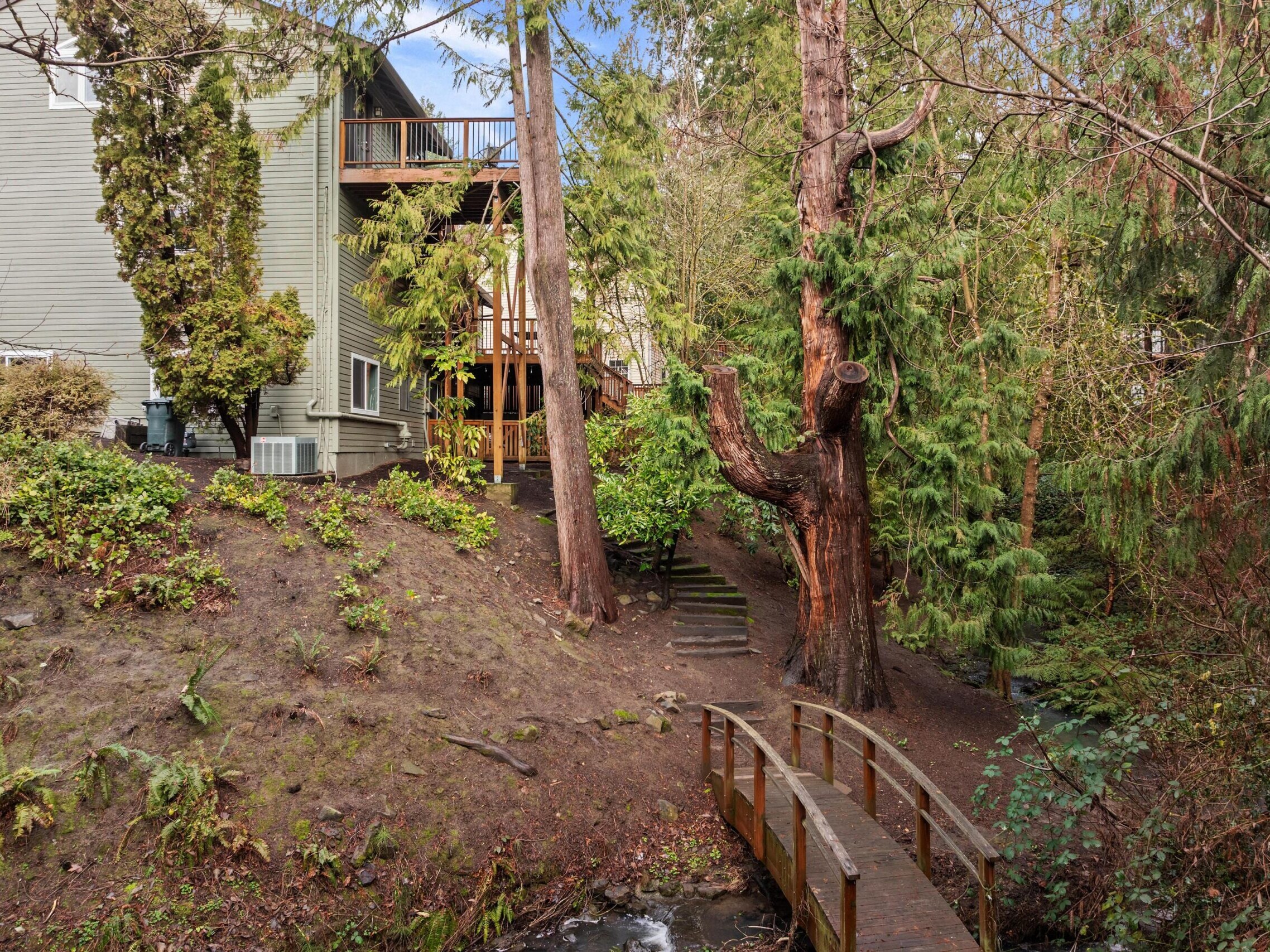 A small wooden footbridge crosses a creek in a forested backyard, leading to a house partially visible on the left. Tall evergreen trees and dense greenery surround the area.