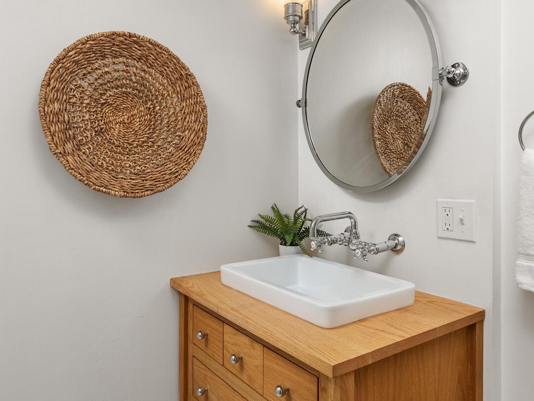 A small bathroom with a wooden vanity, a white vessel sink, a round mirror, two wall-mounted lights, a woven basket wall decor, and a small green plant on the vanity. Towels are neatly stored on the shelf below.