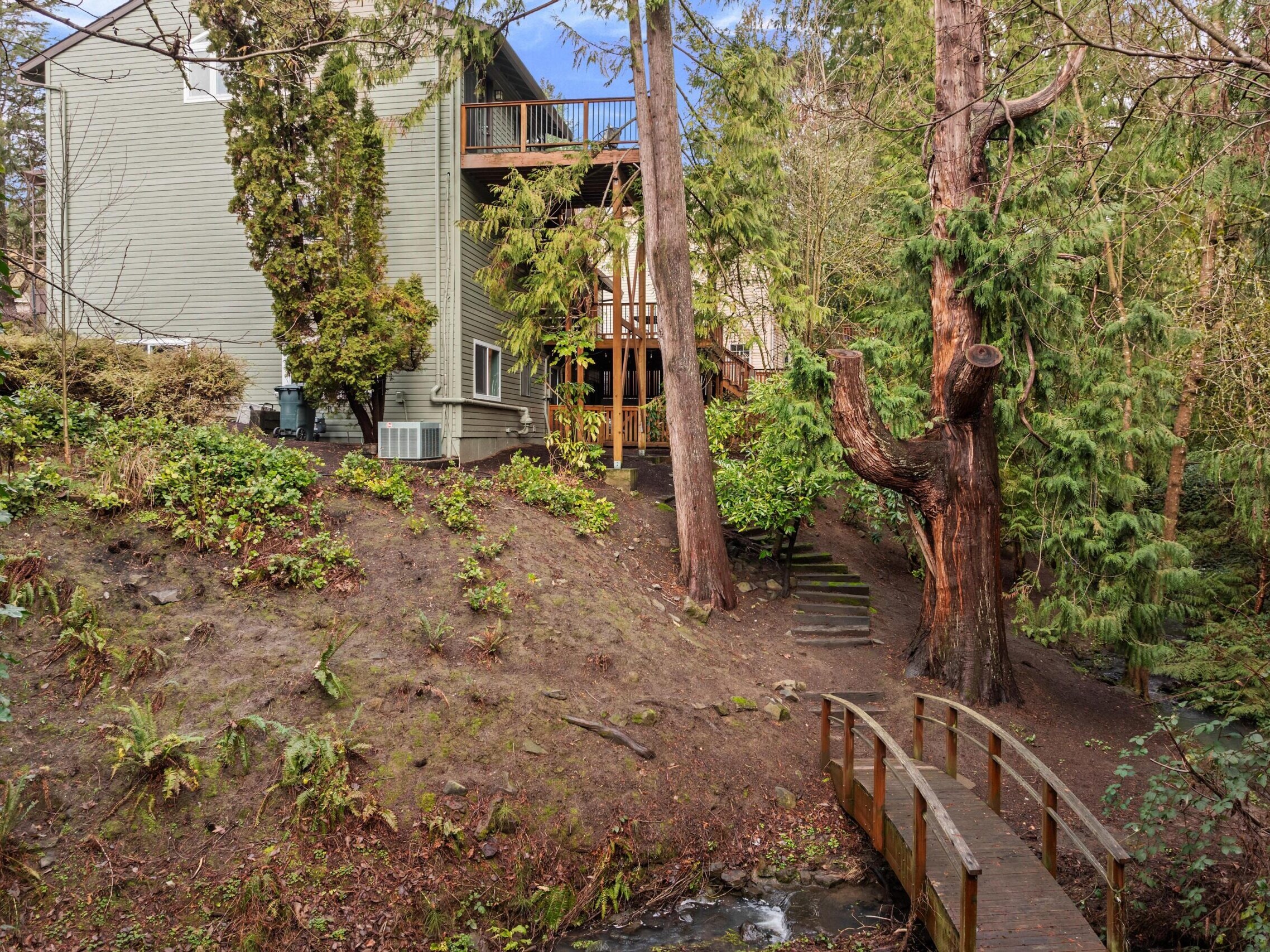 A wooden footbridge crosses a small stream next to a house surrounded by tall trees and lush greenery, with a dirt path and steps leading up to the house.