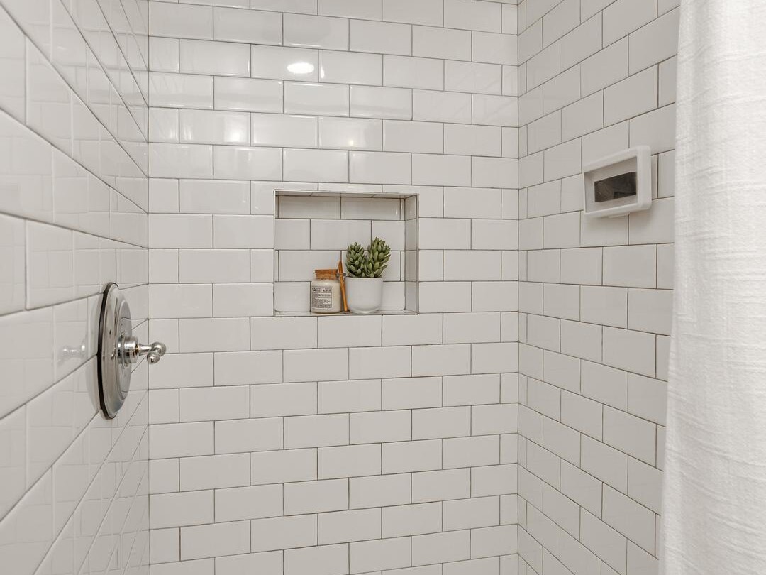 A white-tiled shower with a round metal showerhead, a built-in shelf holding a small potted plant and toiletry items, a silver control knob, and a white shower curtain.