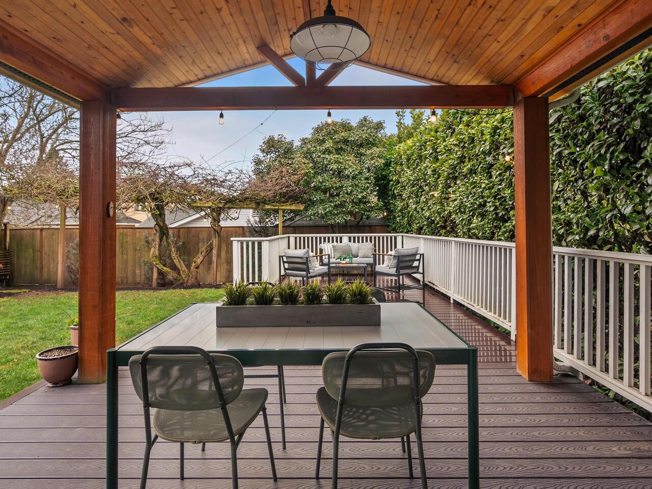 A covered wooden patio with a dining table and chairs overlooks a fenced backyard with grass, trees, and a second outdoor seating area. Tall hedges provide privacy along the white-railed deck.
