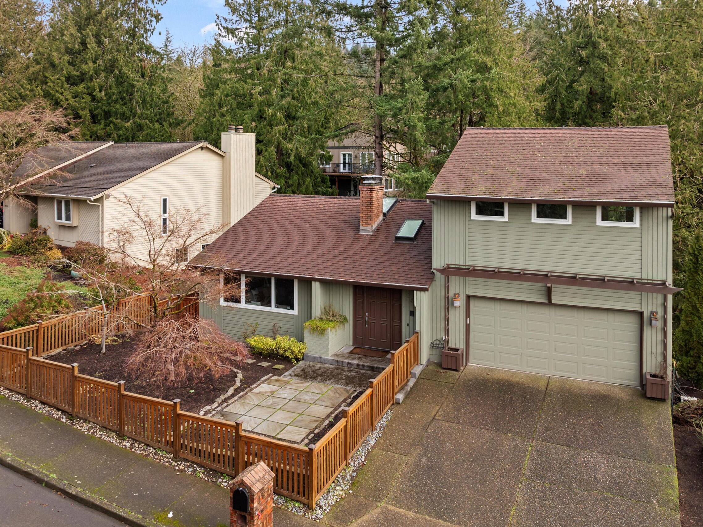 A two-story house with a greenish-gray exterior and brown roof, attached two-car garage, fenced front yard with a small tree, stone walkway, and surrounded by trees and neighboring homes.