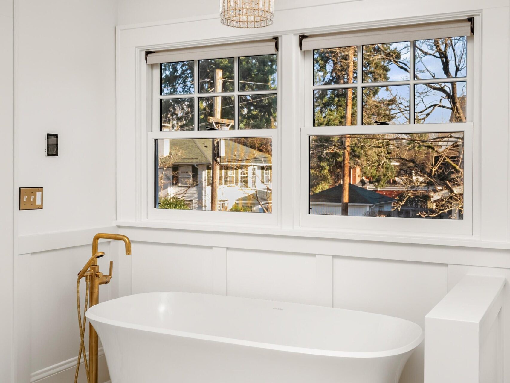 A modern bathroom with a white freestanding tub, gold faucet, herringbone-patterned marble tile floor, white walls, two large windows, and a woven pendant light overhead.