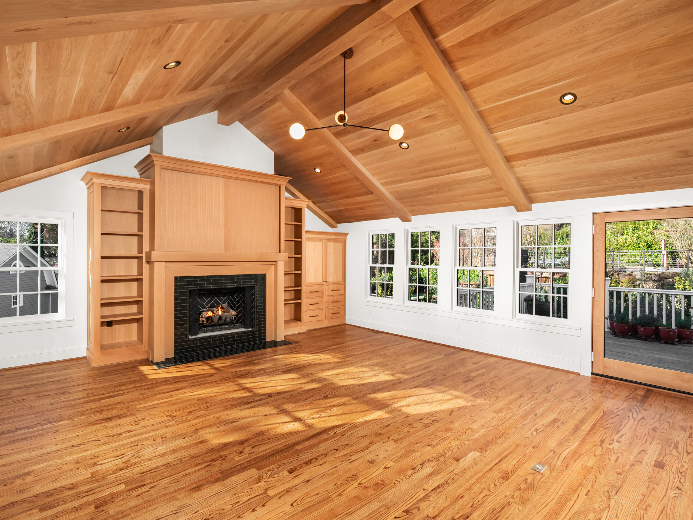 Spacious living room with vaulted wood ceiling, hardwood floors, built-in shelves, fireplace with black tile surround, large windows, and glass door leading to a deck with outdoor views.