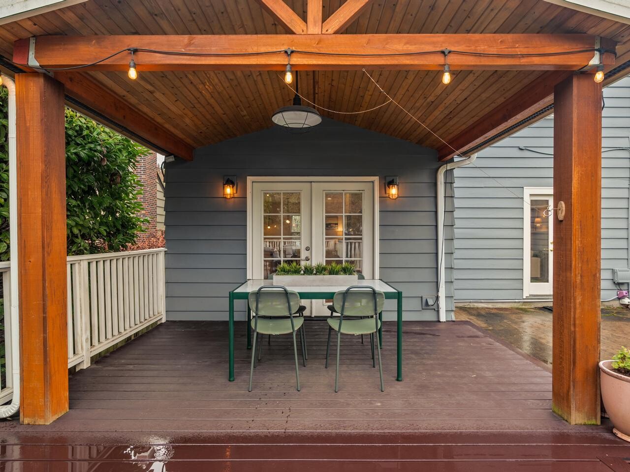 A covered wooden patio with string lights, a glass table, and two chairs, set against a gray house exterior with double windows and a potted plant nearby.
