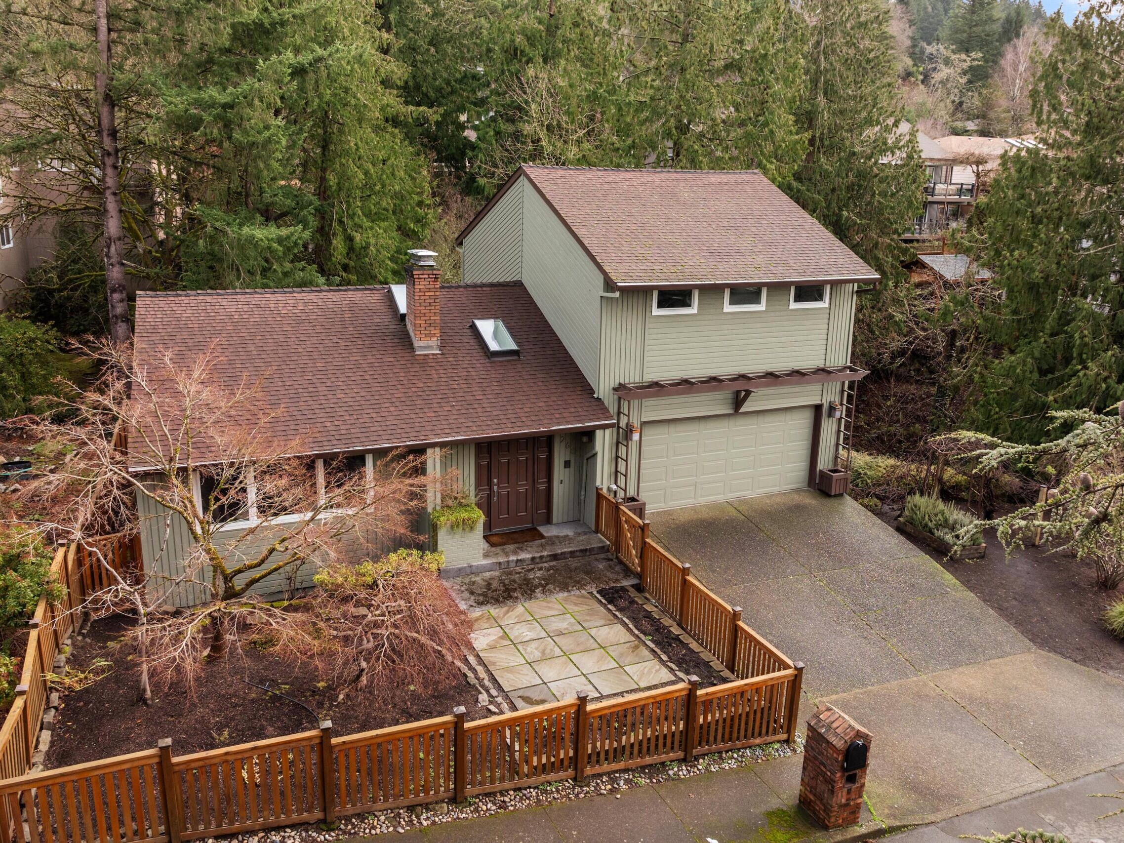 A two-story house with a brown roof, greenish-gray siding, and a two-car garage, surrounded by tall trees and a wooden fence, seen from above. The driveway and front yard are visible.