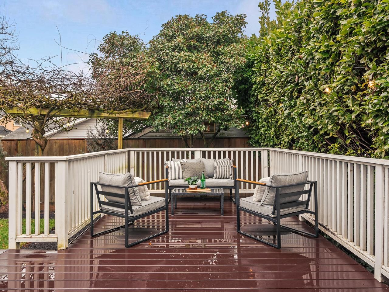 A cozy outdoor patio with four cushioned chairs around a small table on a wet wooden deck, surrounded by white railings and lush green trees and bushes.