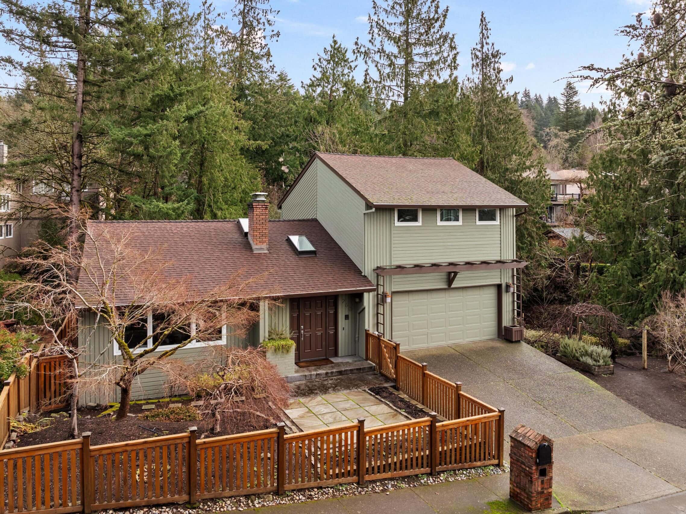 Two-story house with green siding, brown roof, attached double garage, and a wooden fence surrounding the front yard. The house is set among tall trees and has a stone pathway leading to the front door.