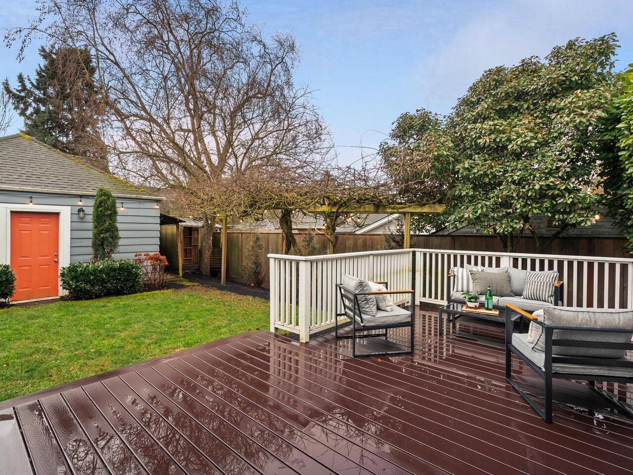A backyard with a wooden deck featuring outdoor seating, cushions, and a small table. The deck overlooks a grassy lawn, trees, shrubs, and a gray shed with an orange door. The sky is partly cloudy.